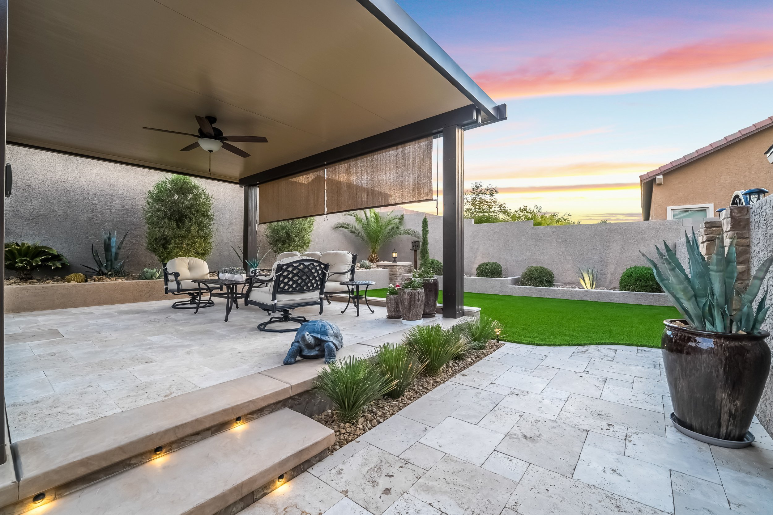 Backyard patio with tiled flooring, outdoor furniture, potted plants, a turtle statue, a grassy area, and a sunset sky overhead.