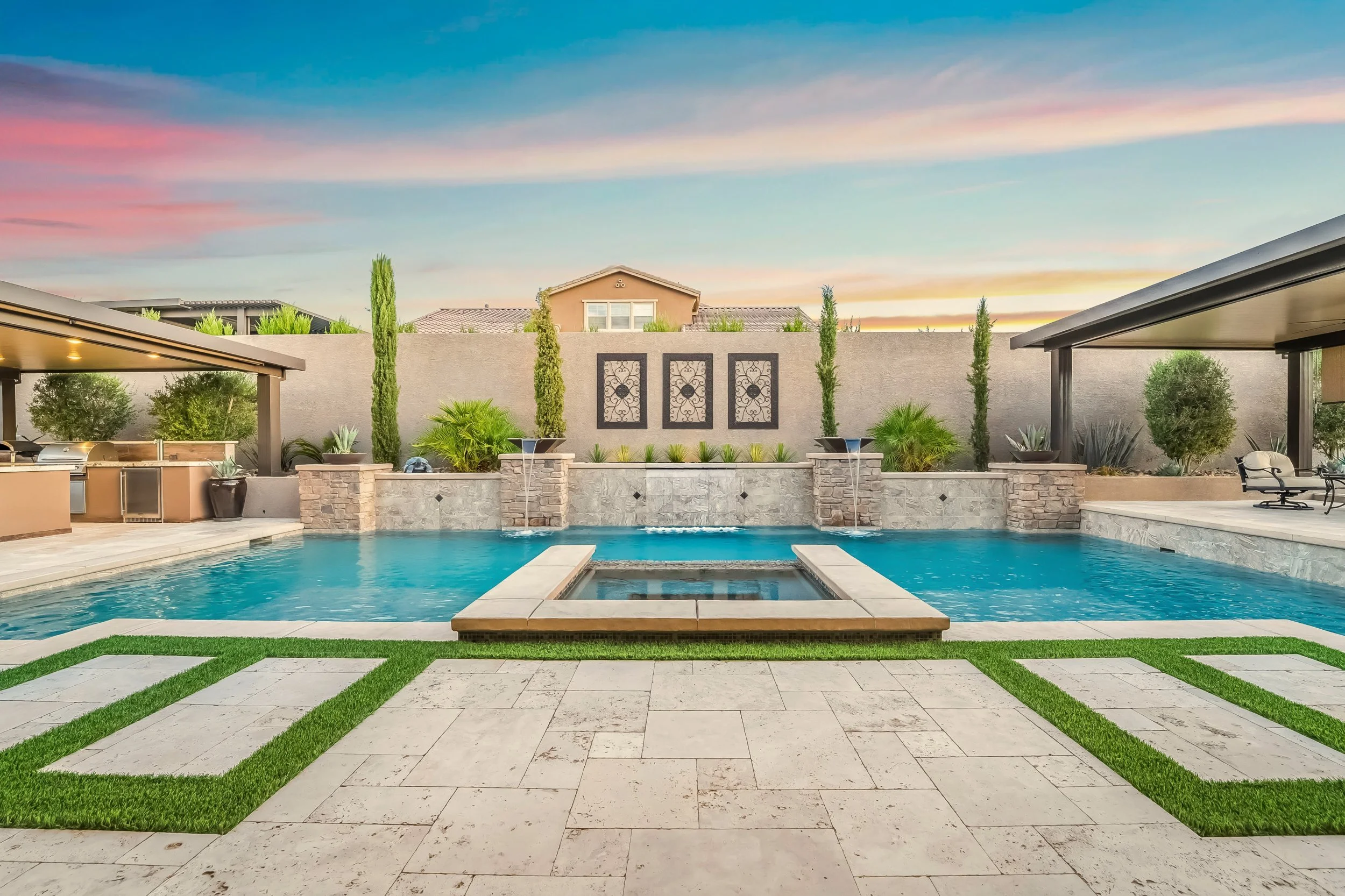 Backyard with a swimming pool, stone waterfall, surrounded by potted plants, tall trees, and a seating area under the patio, sunset sky