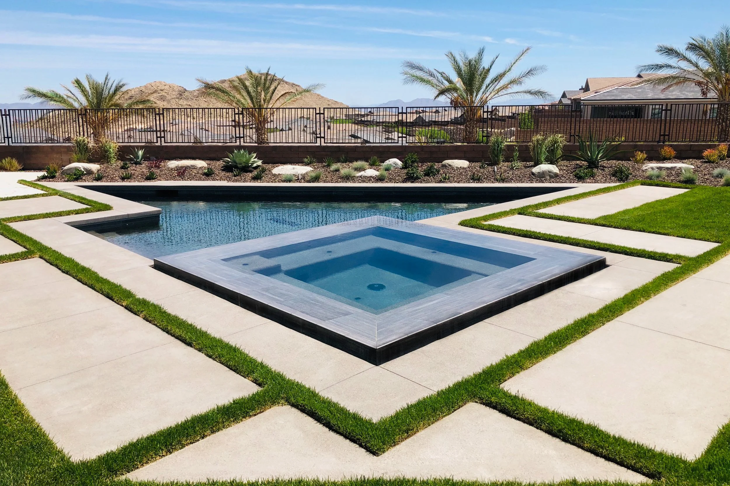 A backyard with a modern swimming pool and spa, surrounded by green grass and stone pathways. There are desert plants, palm trees, and a mountain view in the background.