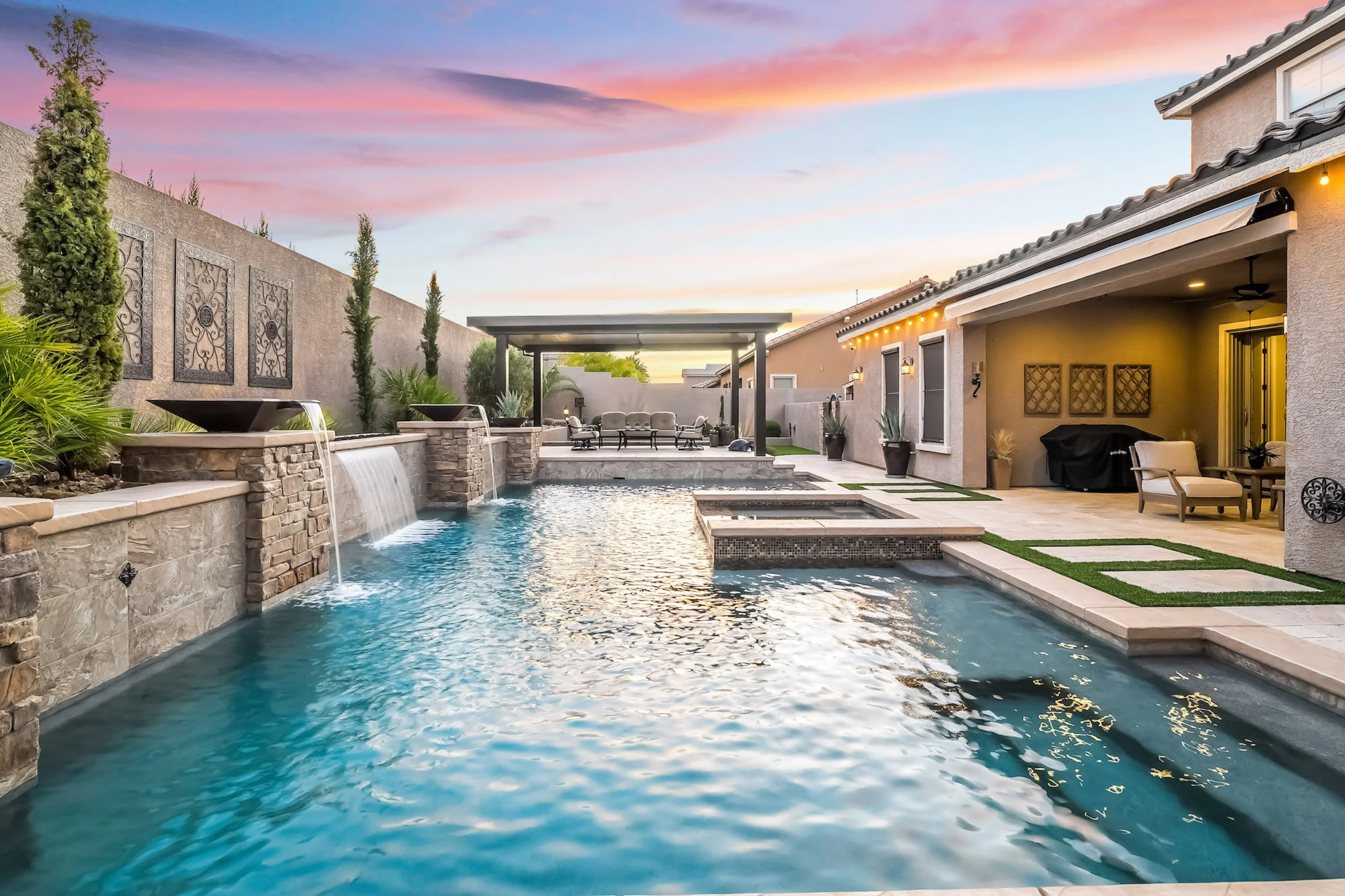 A backyard swimming pool area during sunset with a covered seating area, patio furniture, and water features.
