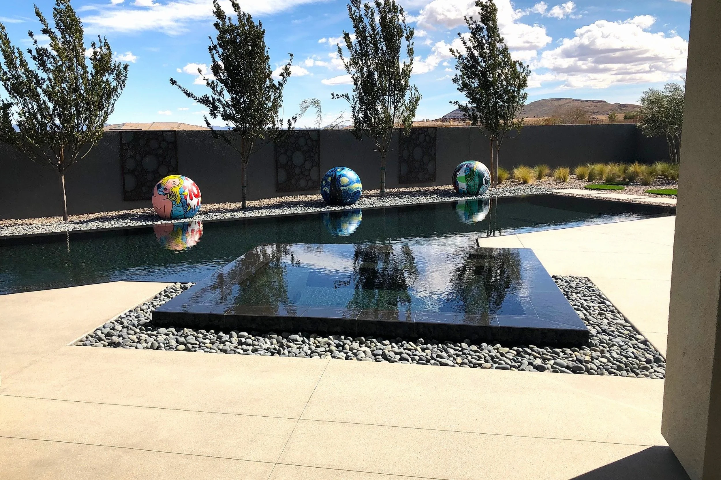 Modern backyard with a rectangular reflecting pool, three painted spheres, small trees, and a gray wall with decorative panels.