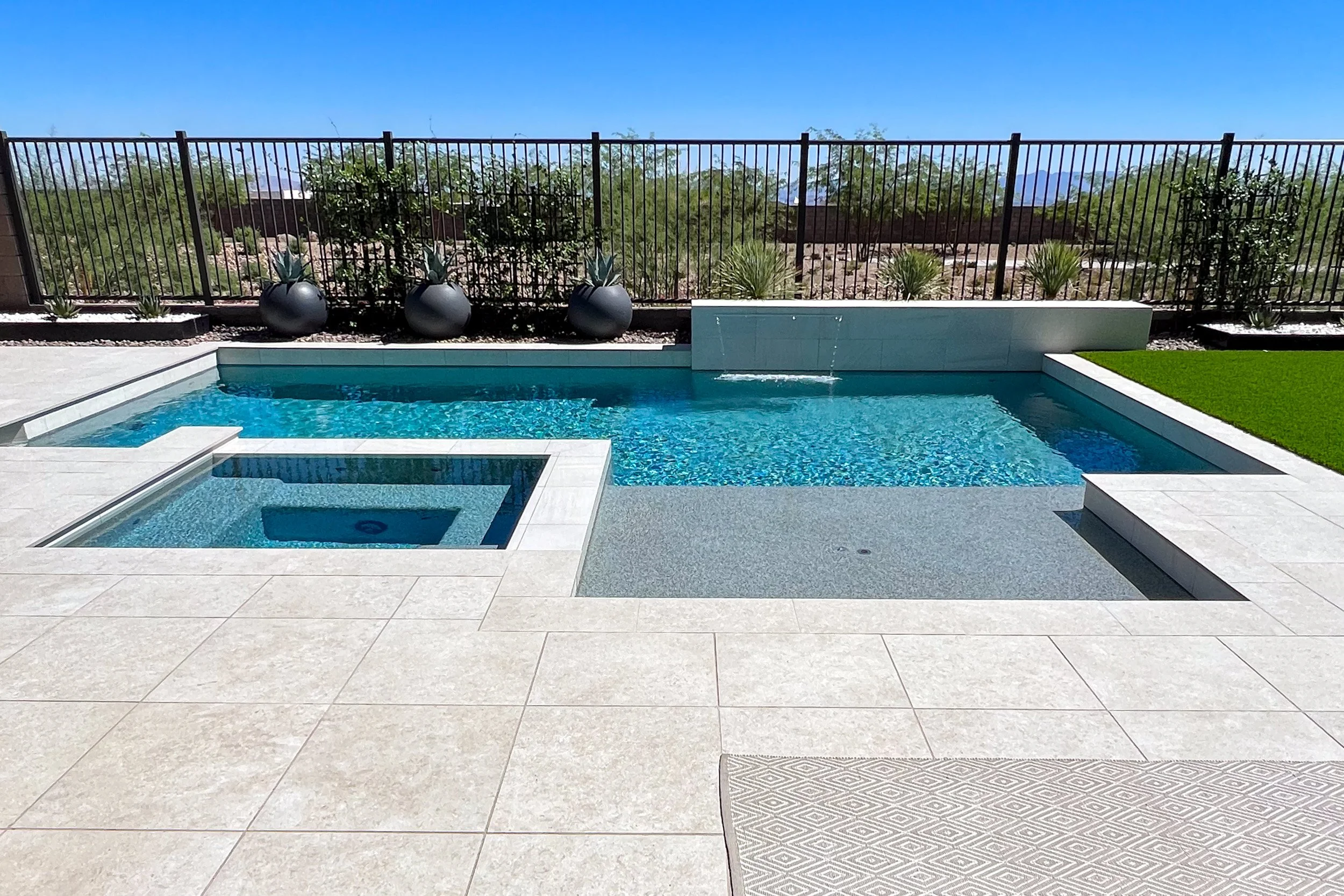 A modern backyard swimming pool with a hot tub, surrounded by beige tiled patio, a black fence, desert landscape, and mountains in the background.