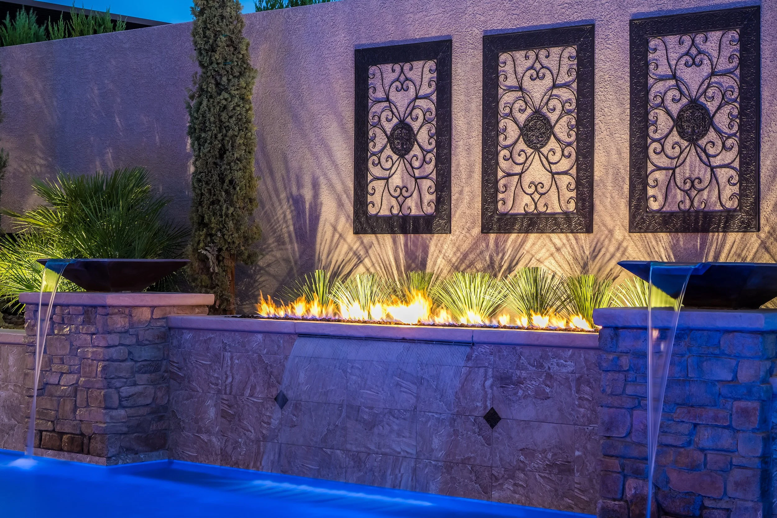 Outdoor water feature with lit fire and stone wall background, three ornate metal wall art pieces, two black water bowls with water flowing into a pool.
