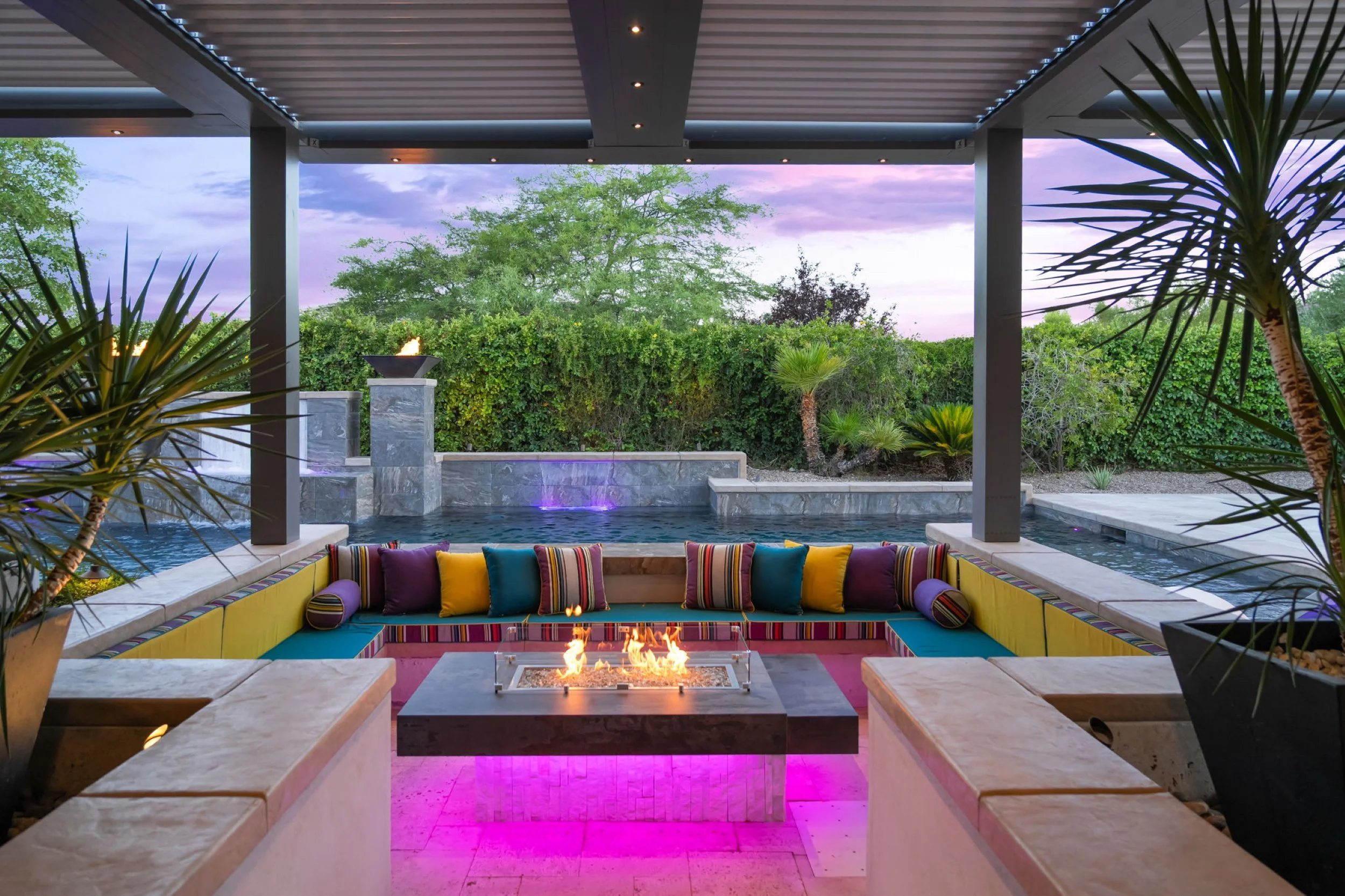 Outdoor patio with colorful seating around a fire pit, overlooking a pool and lush greenery at sunset.