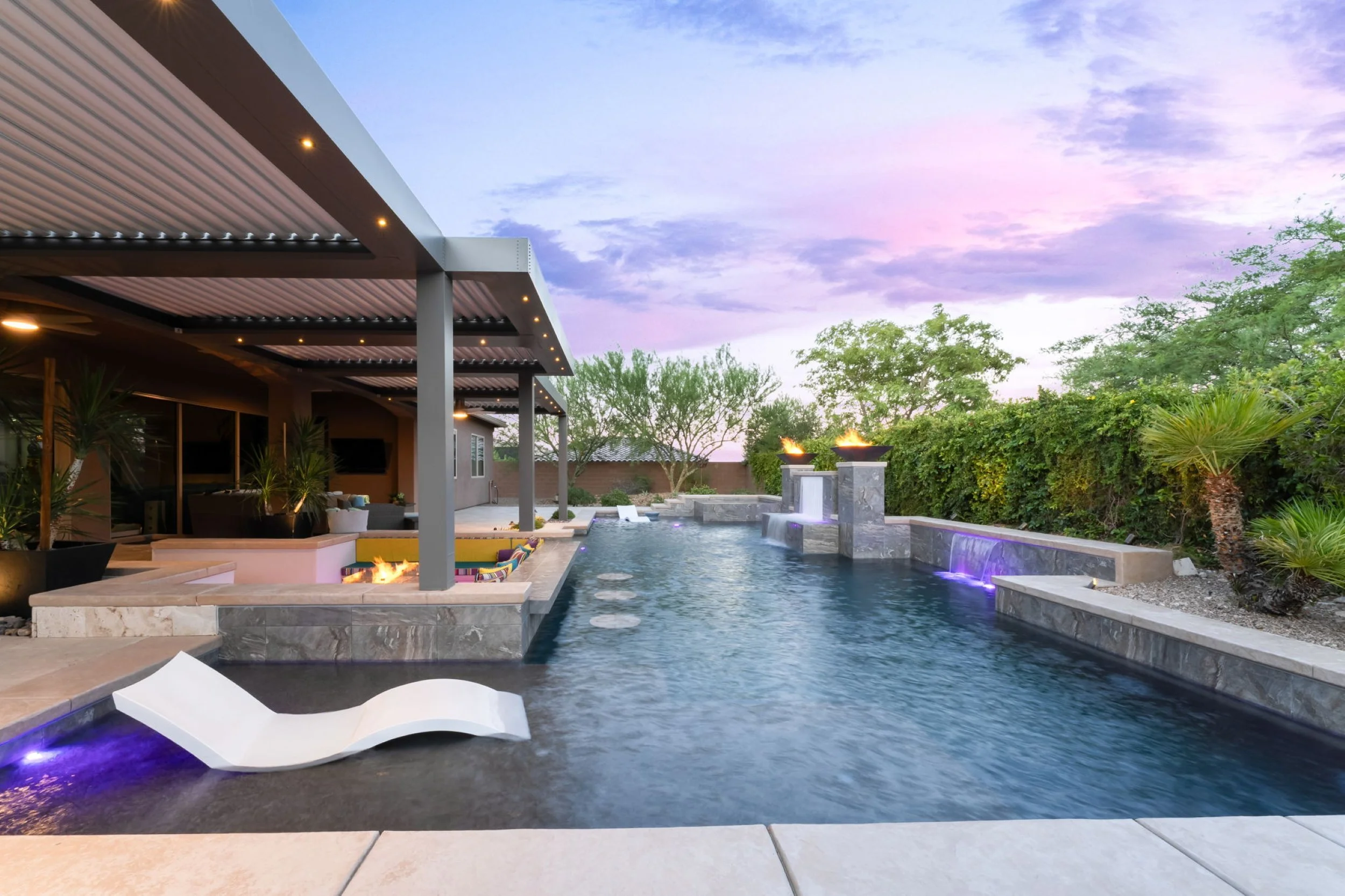 Backyard with a modern swimming pool, waterfalls, fire pits, lounge chairs, and outdoor seating area during twilight.