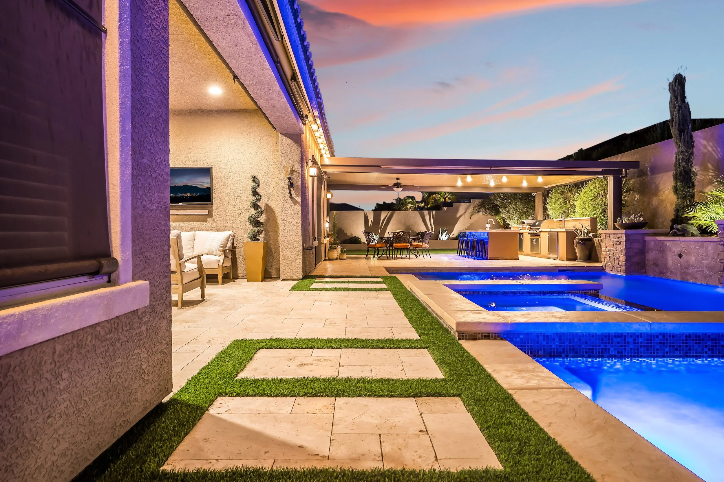 A backyard with a swimming pool, outdoor dining area, and outdoor kitchen at sunset. The pool area has blue lighting, and there is modern patio furniture and potted plants.