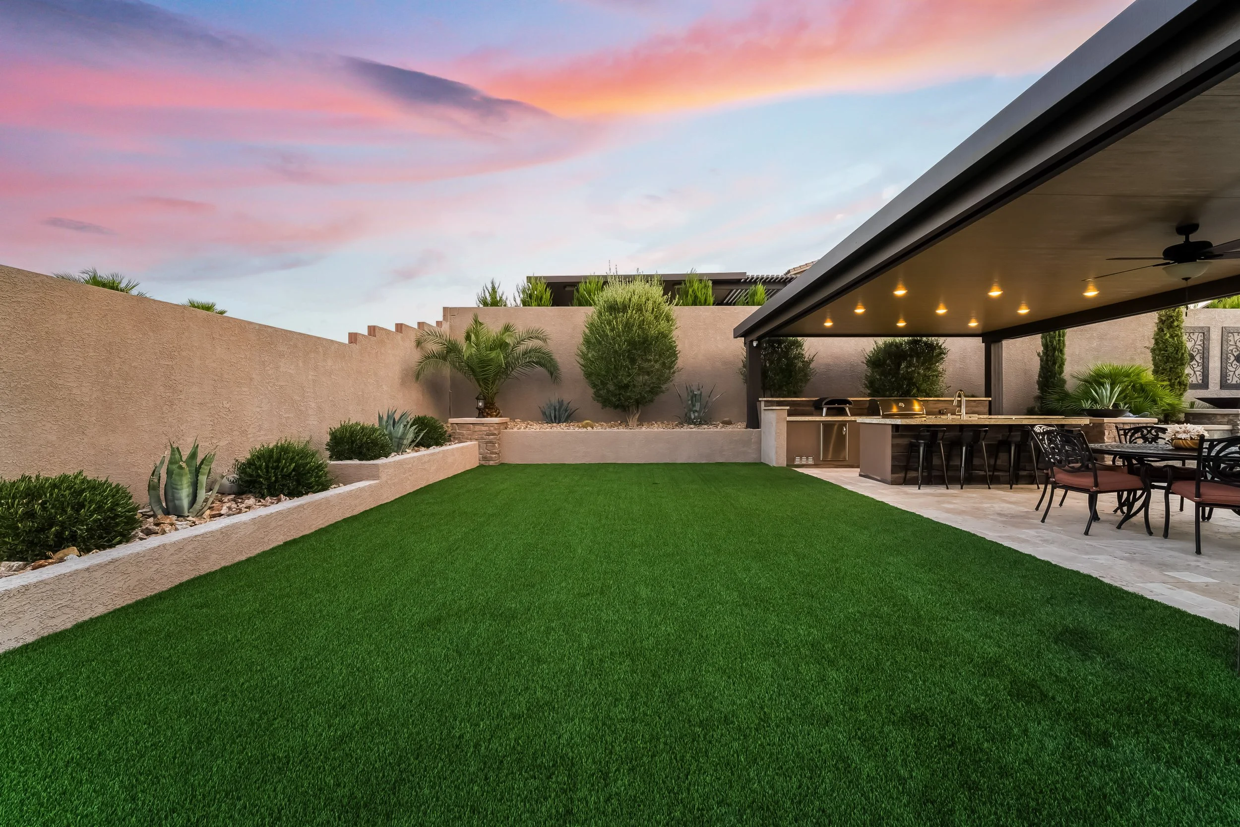 Backyard with manicured green lawn, desert-style plants, and an outdoor kitchen and dining area under a covered patio at sunset.