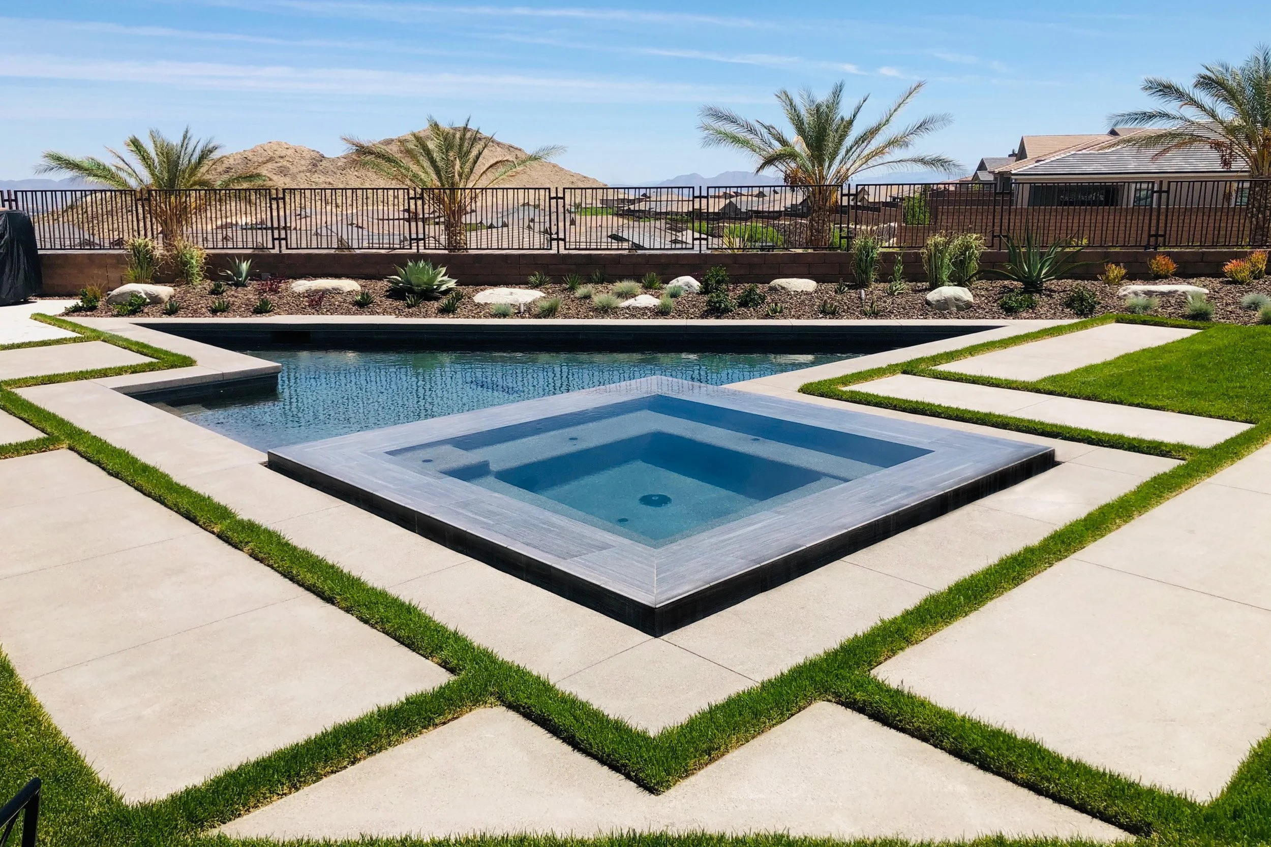 A backyard with a modern swimming pool and spa, surrounded by green grass and stone pathways. There are desert plants, palm trees, and a mountain view in the background.