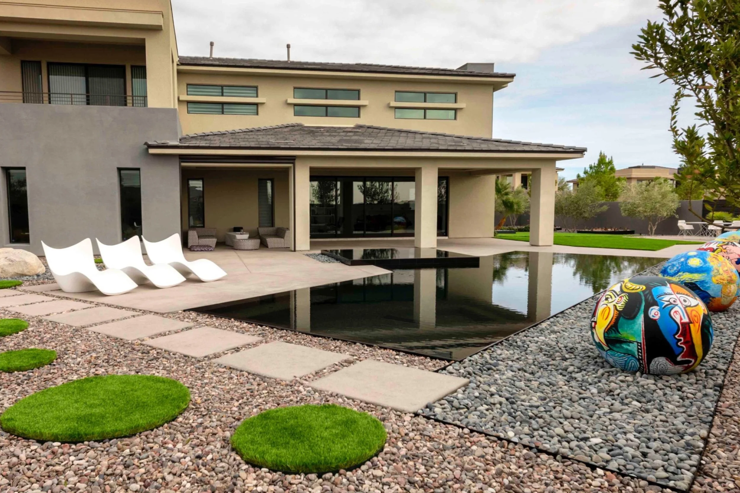 Modern house with a backyard featuring a reflective black pool, white lounge chairs, colorful decorative spheres, green grass patches, tiled walkway, and outdoor furniture.