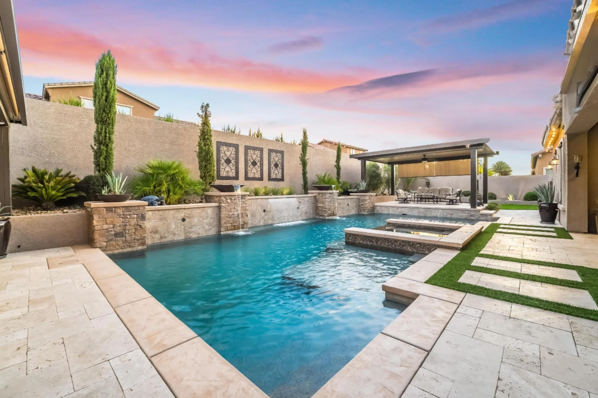 Backyard with a rectangular swimming pool, stone waterfall features, outdoor furniture under a covered patio, landscaped with plants and tall trees, and a colorful sunset sky.