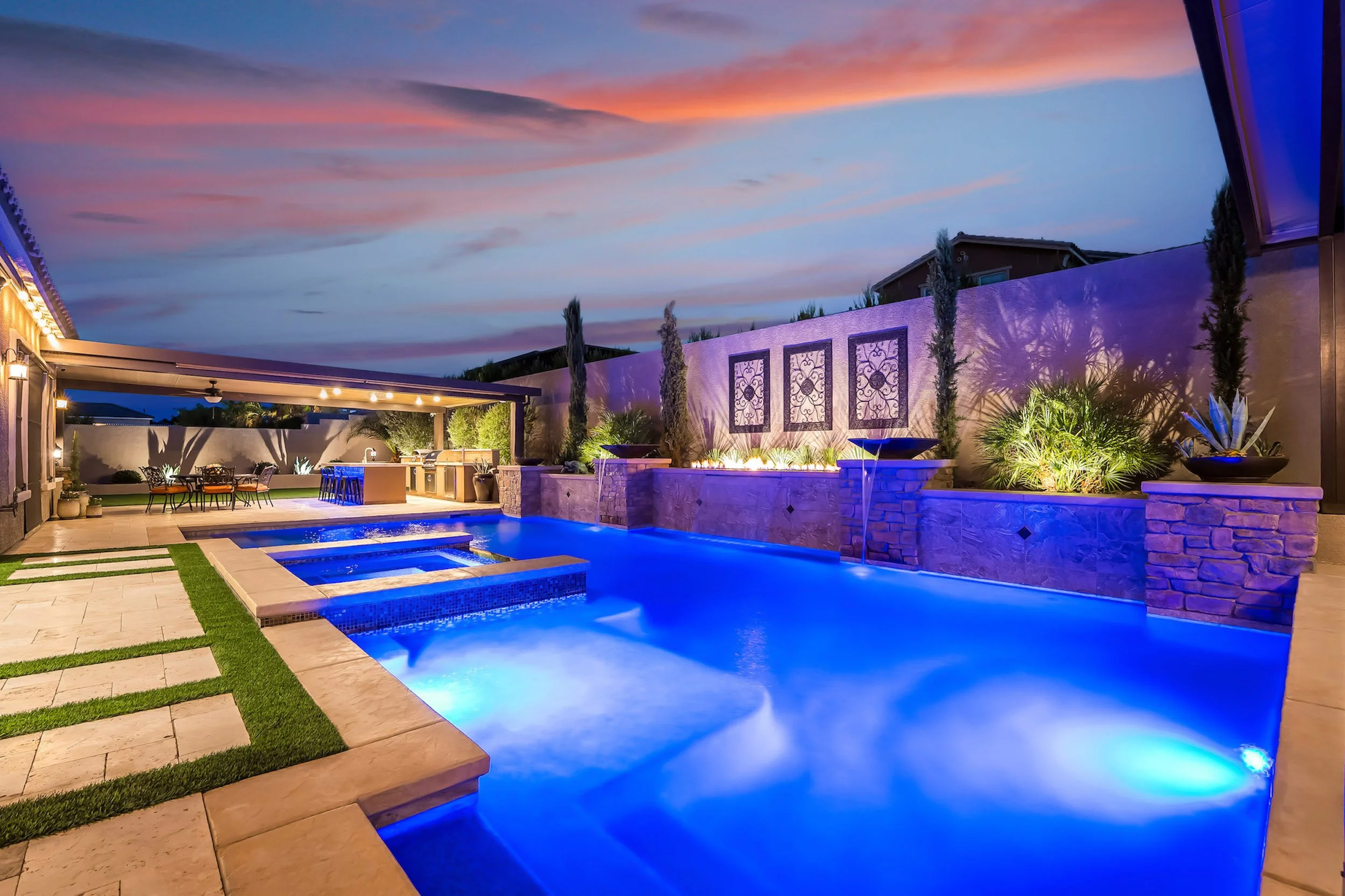Backyard at sunset with illuminated swimming pool, outdoor dining area, lush plants, and decorative wall art.
