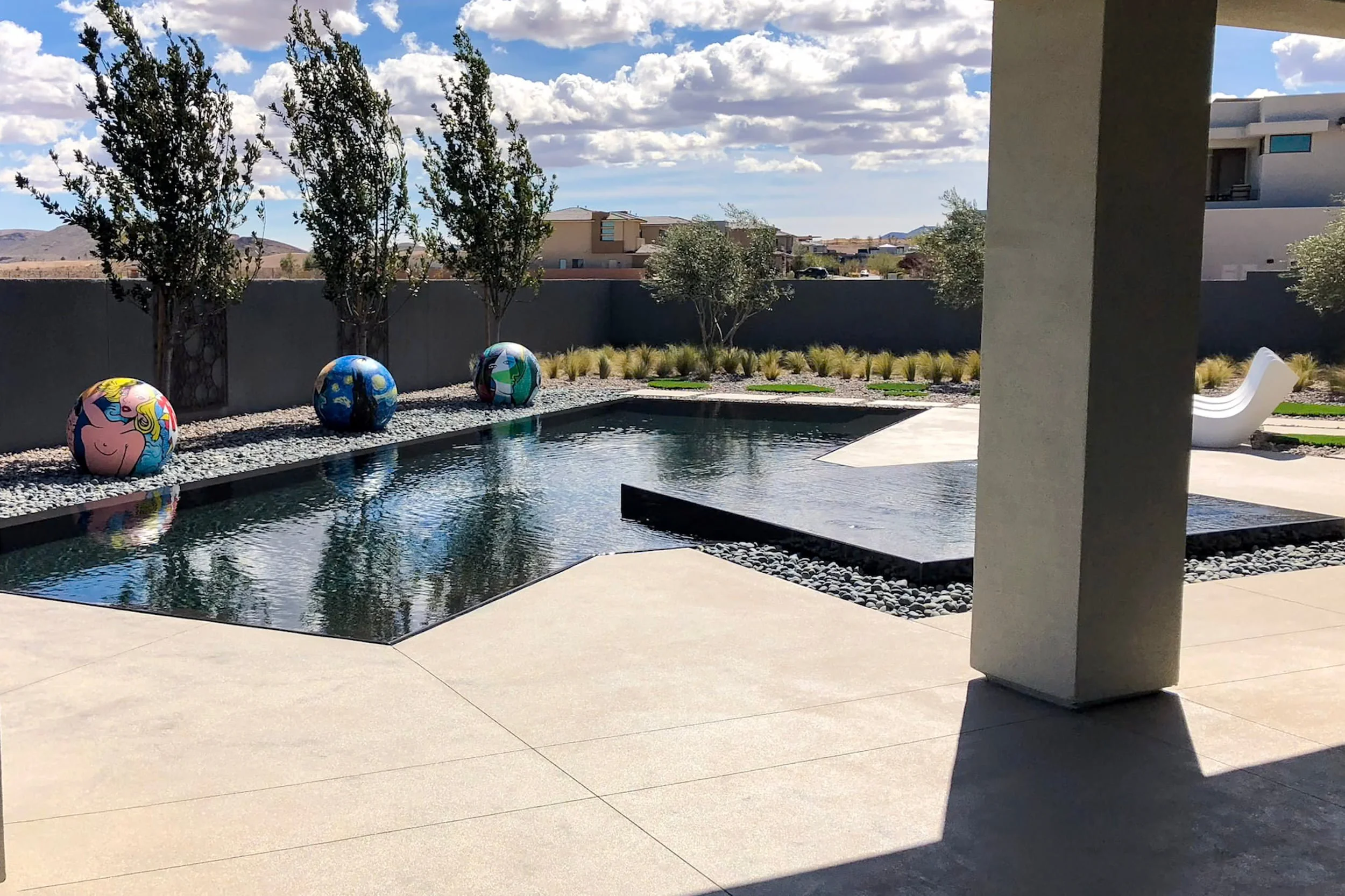 A modern backyard with a rectangular swimming pool, decorative painted spheres, trees, gravel, ornamental grasses, and a patio with a lounge chair under a covered area.