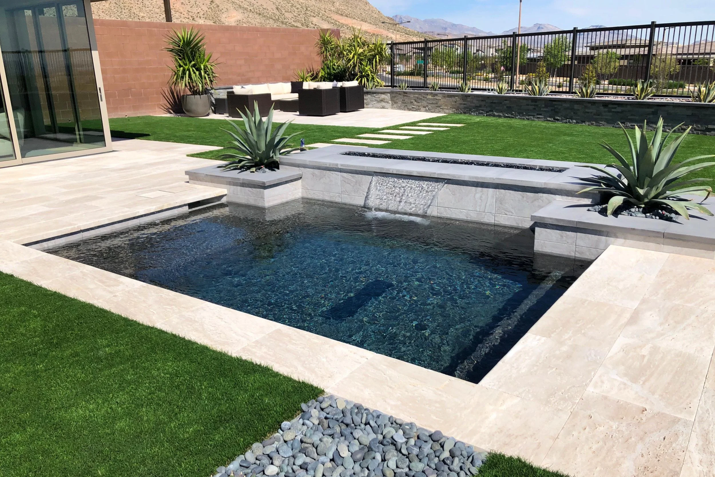 Modern backyard with a rectangular swimming pool, surrounded by stone tiles, green grass, and desert plants. There is a water feature flowing into the pool, and an outdoor seating area with black furniture and white cushions in the background. A glas