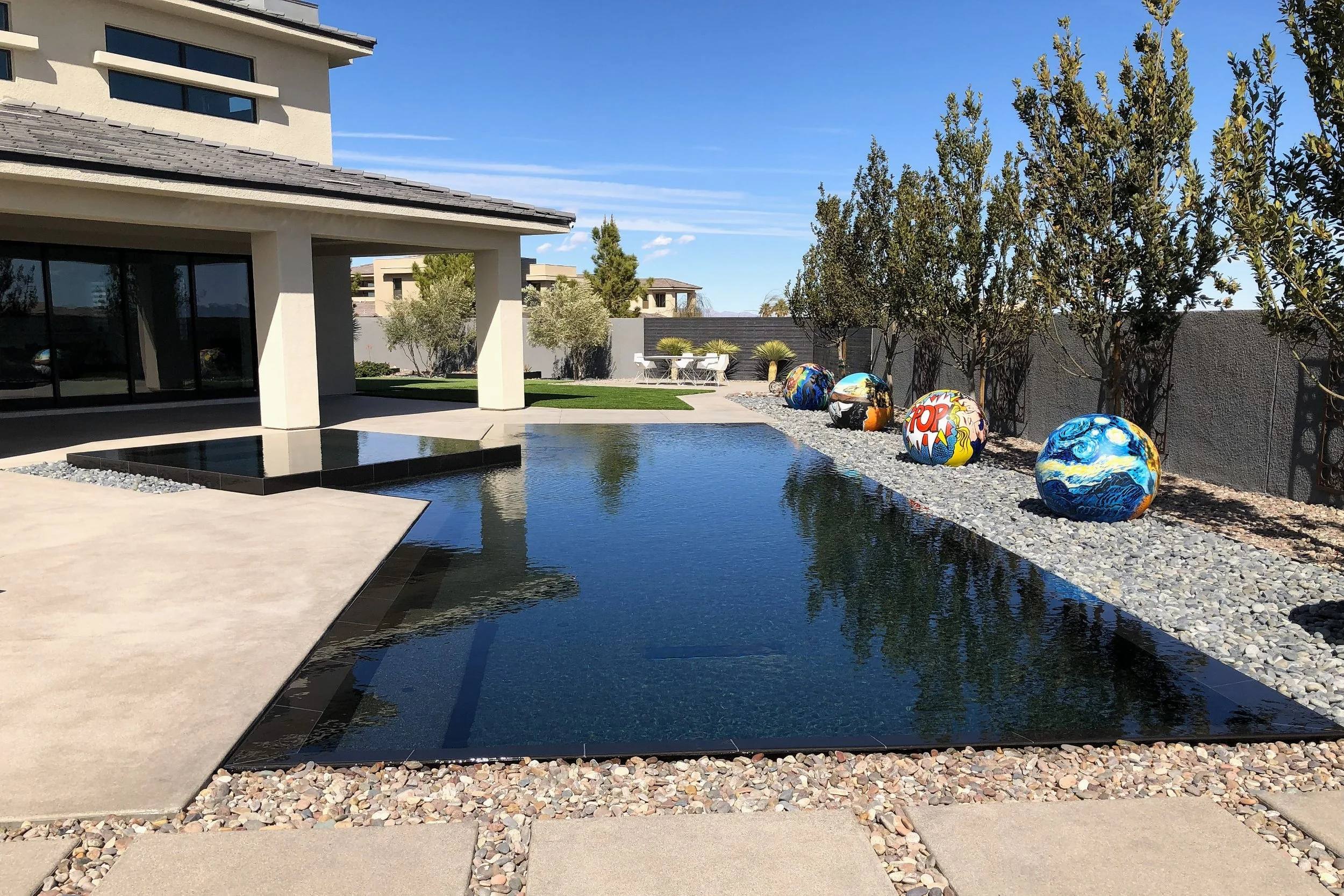 A modern backyard with a rectangular reflective pool, colorful painted rocks resembling artwork, trees, and outdoor seating under a blue sky.