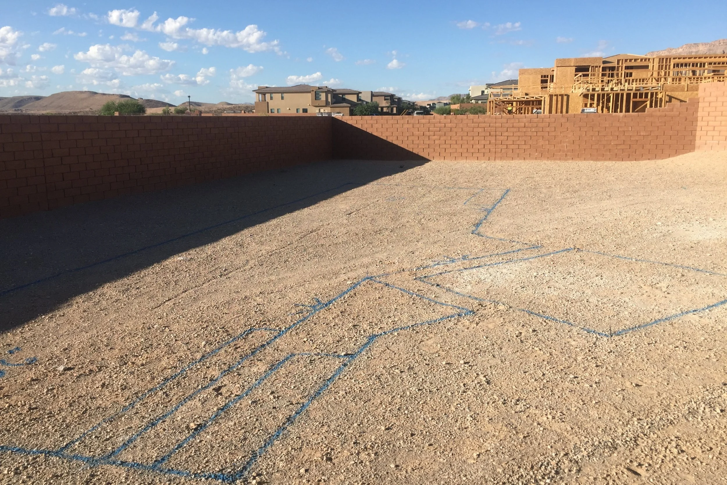 A construction site with a gravel ground, blue chalk hopscotch outlines, a brick wall, and multiple houses under construction in the background under a partly cloudy sky.