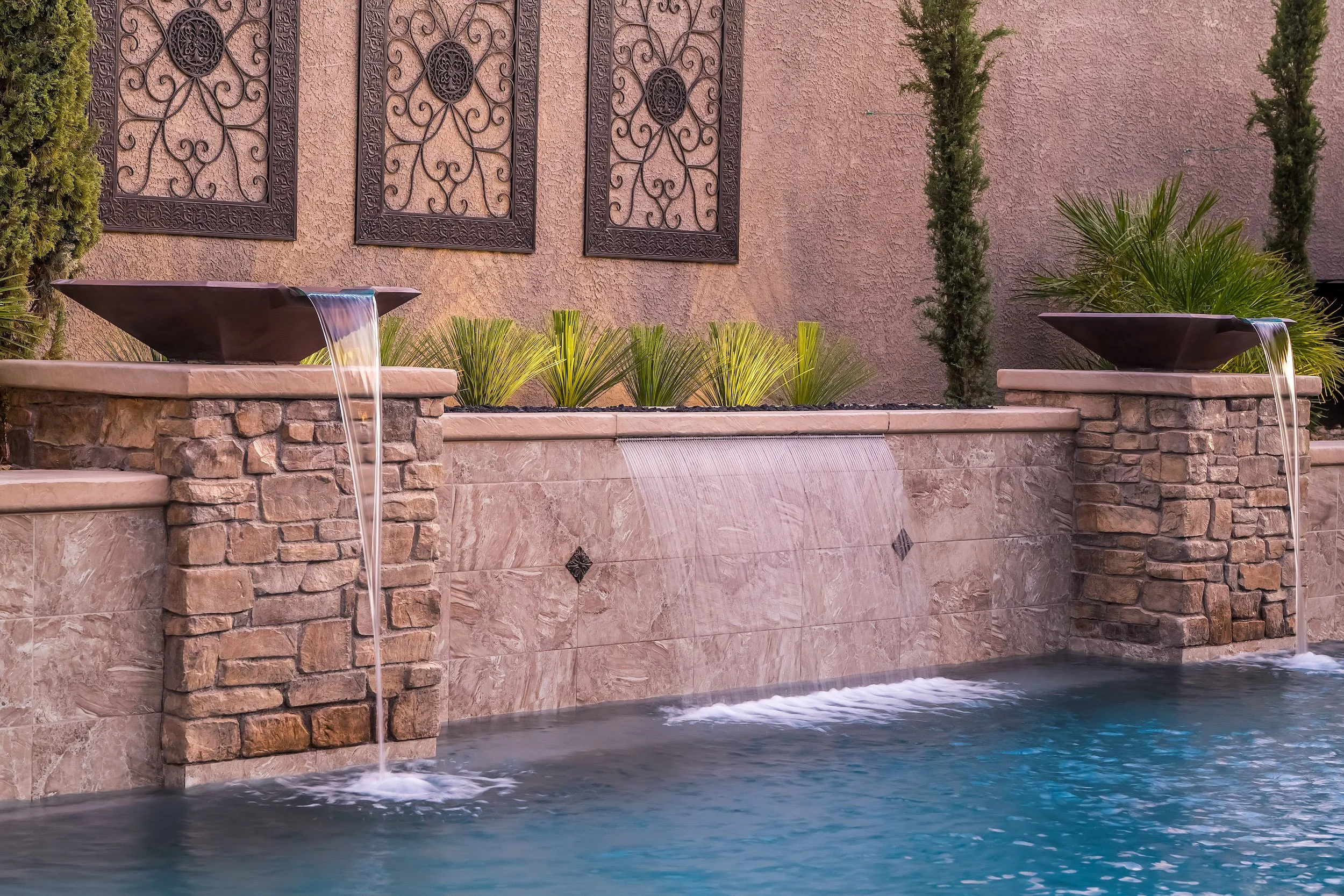 A decorative water fountain feature with two rectangular black bowls on stone pedestals, water flowing from each into a pool, with a pink stucco wall with metal wall decorations, plants, and trees in the background.