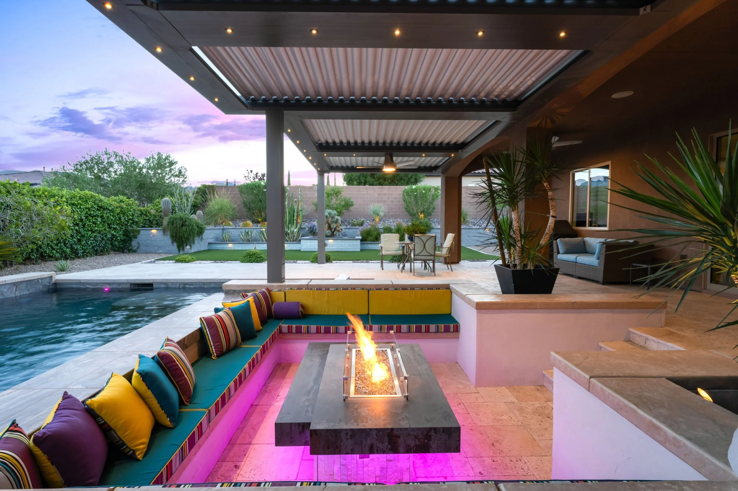 Modern outdoor patio with colorful cushioned seating around a fire pit, adjacent to a swimming pool, with a dining table and chairs, lush desert landscaping, and a purple sunset sky in the background.