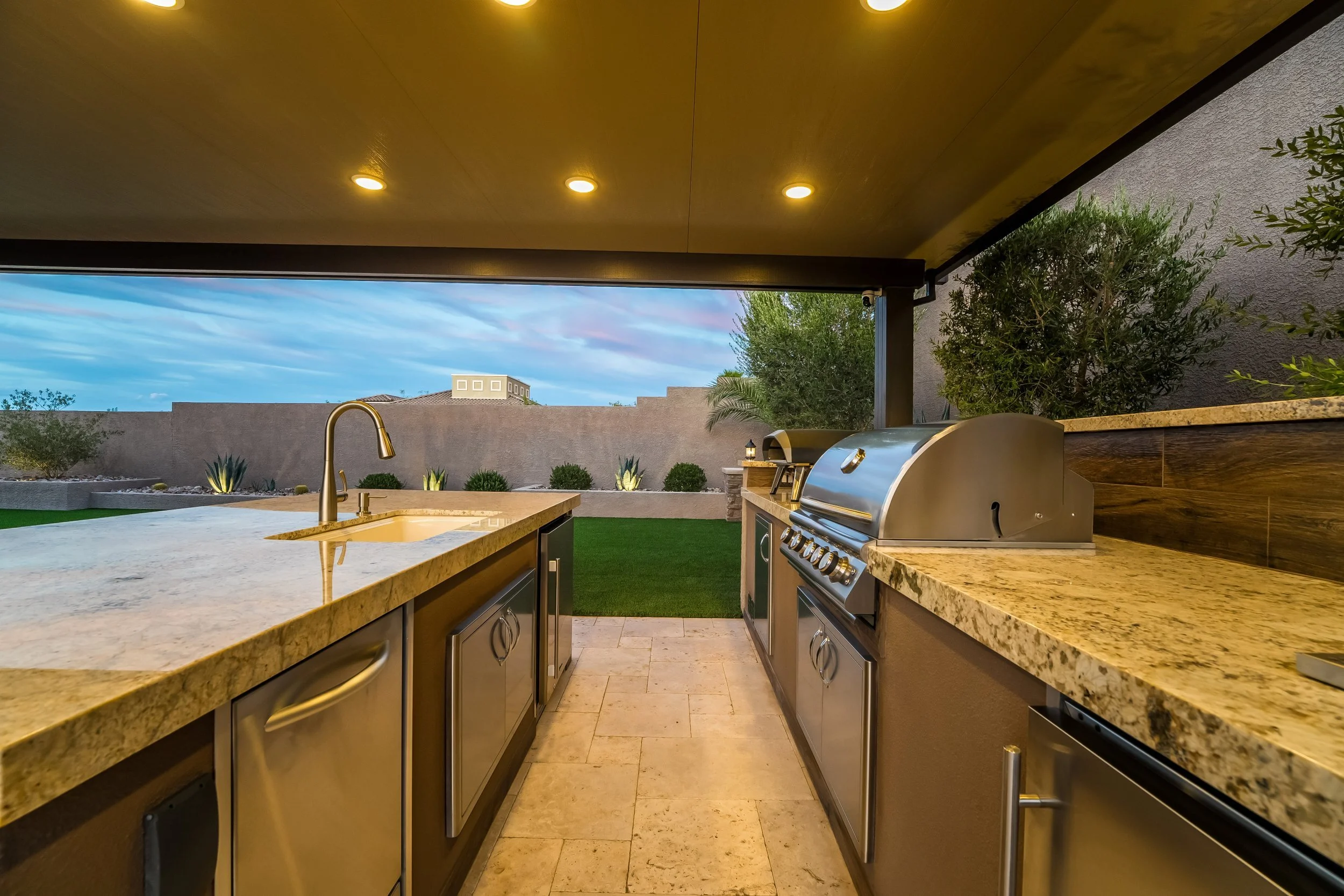 Outdoor kitchen with granite countertops, stainless steel appliances, and a built-in grill, overlooking a backyard with a grass lawn and decorative plants, under a covered patio with ceiling lights.