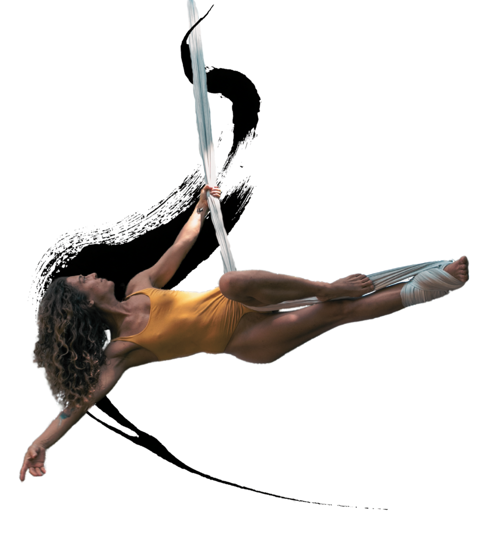 A woman with curly hair in a yellow leotard performing an aerial silk routine, hanging horizontally with one hand holding the silk and legs extended.