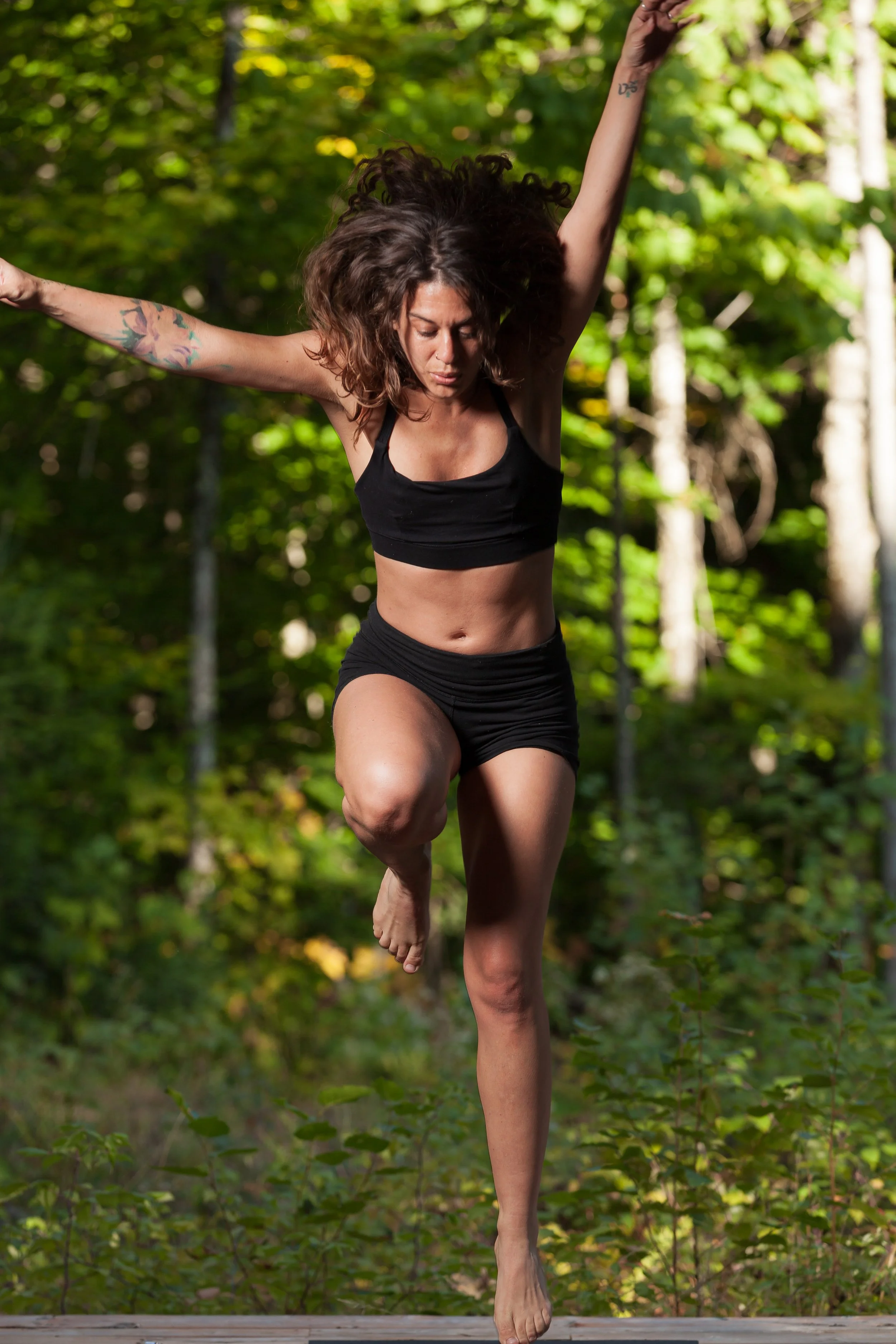 A woman practicing yoga outdoors in a forest, balancing on one leg with her arms raised.