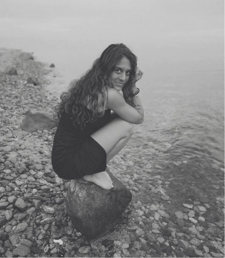 A woman with long curly hair squatting on a rock at the edge of a pebble beach near a body of water, looking at the camera.