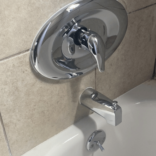 Close-up of a chrome shower faucet and handle with a bathtub below, in a tiled bathroom.