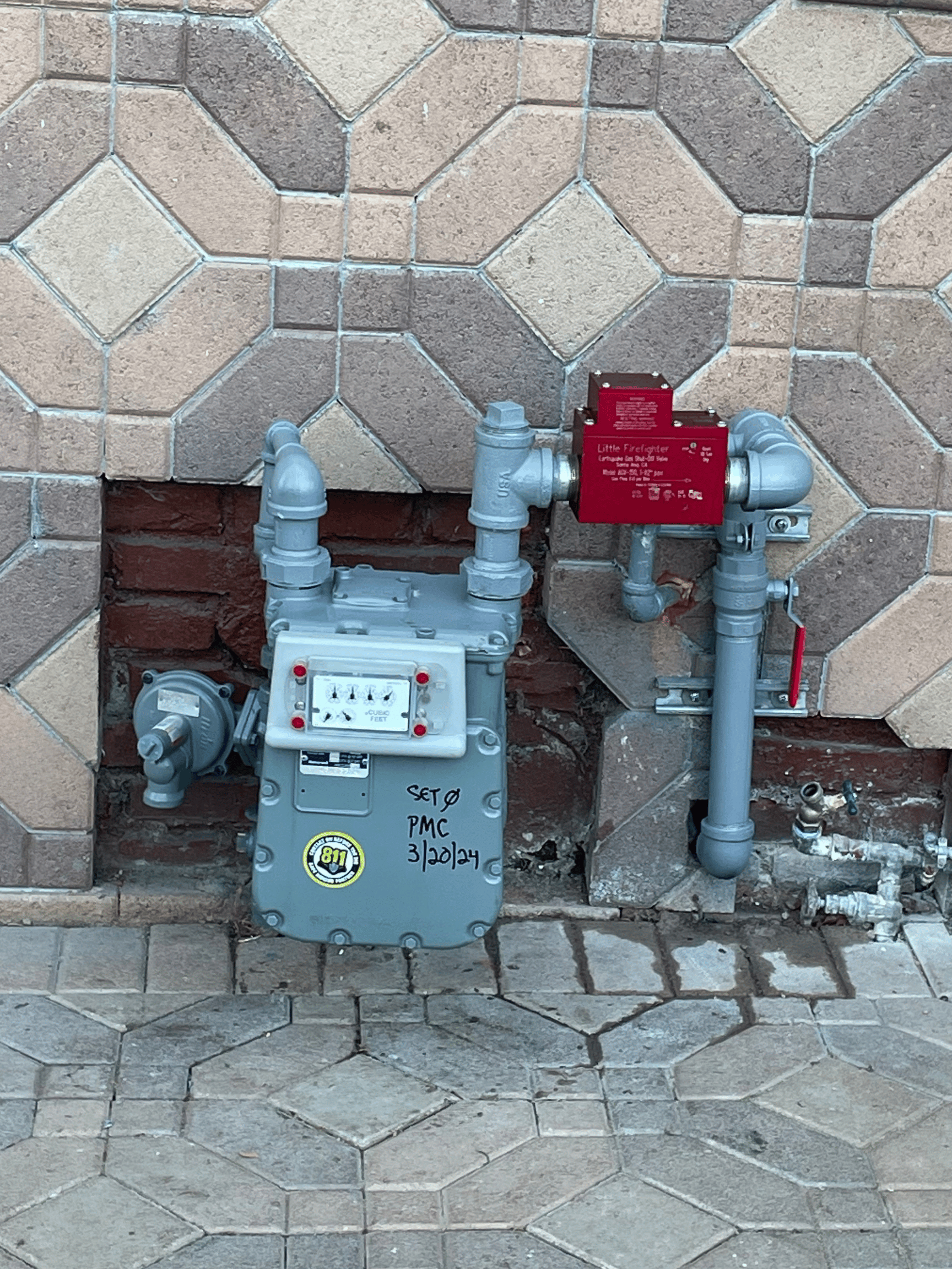 Plumber repairing an outdoor gas line near a gas meter, using pliers and wearing safety gloves, with pipe fittings and tools organized in a red tray beside him.
