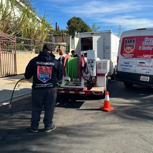Chavez plumbing technician performing hydro jetting by feeding a high-pressure sewer jetter hose into an outdoor cleanout, with a trailer mounted jetter and branded service van in the background
