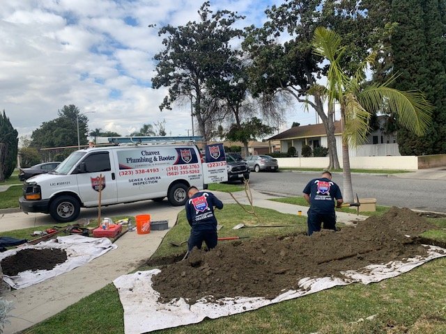 Two workers digging in a yard near a sidewalk with a Chavez Plumbing & Router van parked nearby, under a partly cloudy sky with trees and houses in the background.