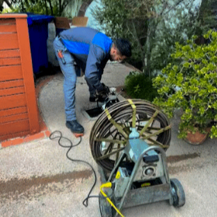 A man is operating a pressure washer on a concrete driveway next to a wooden fence and potted plants.