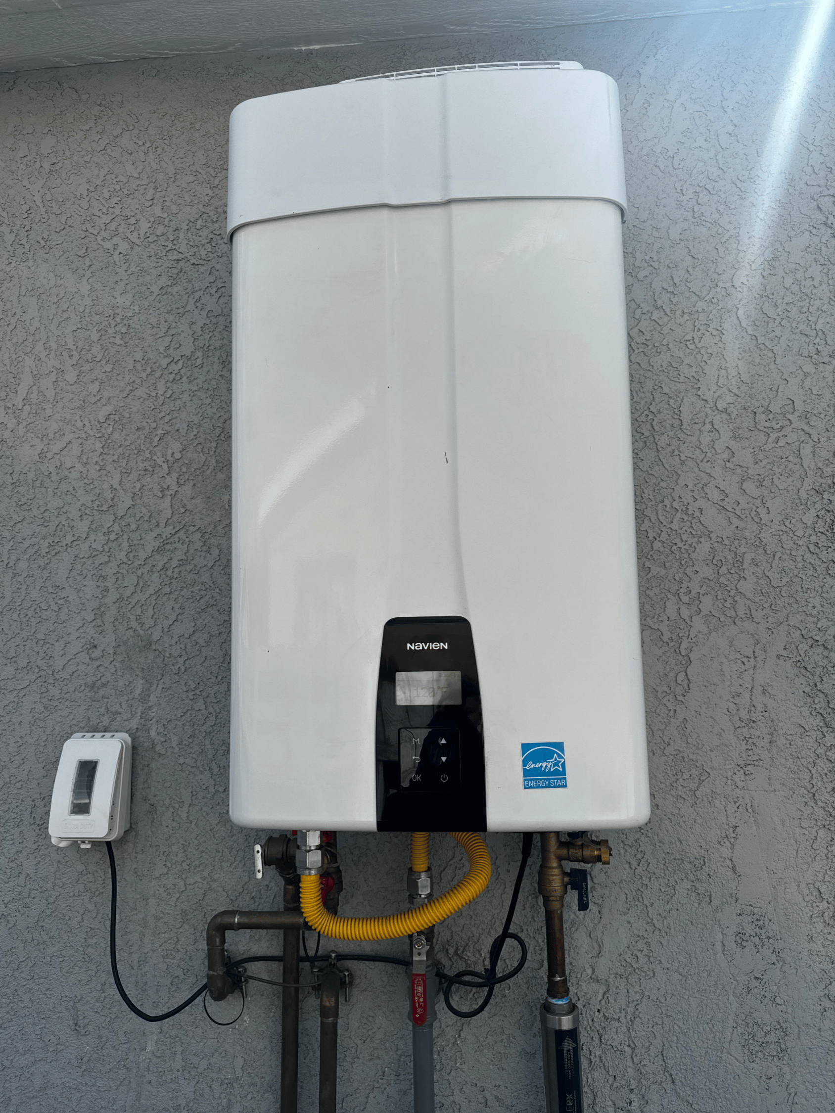 An outdoor white tankless water heater mounted on a stucco wall with pipes and connectors below.