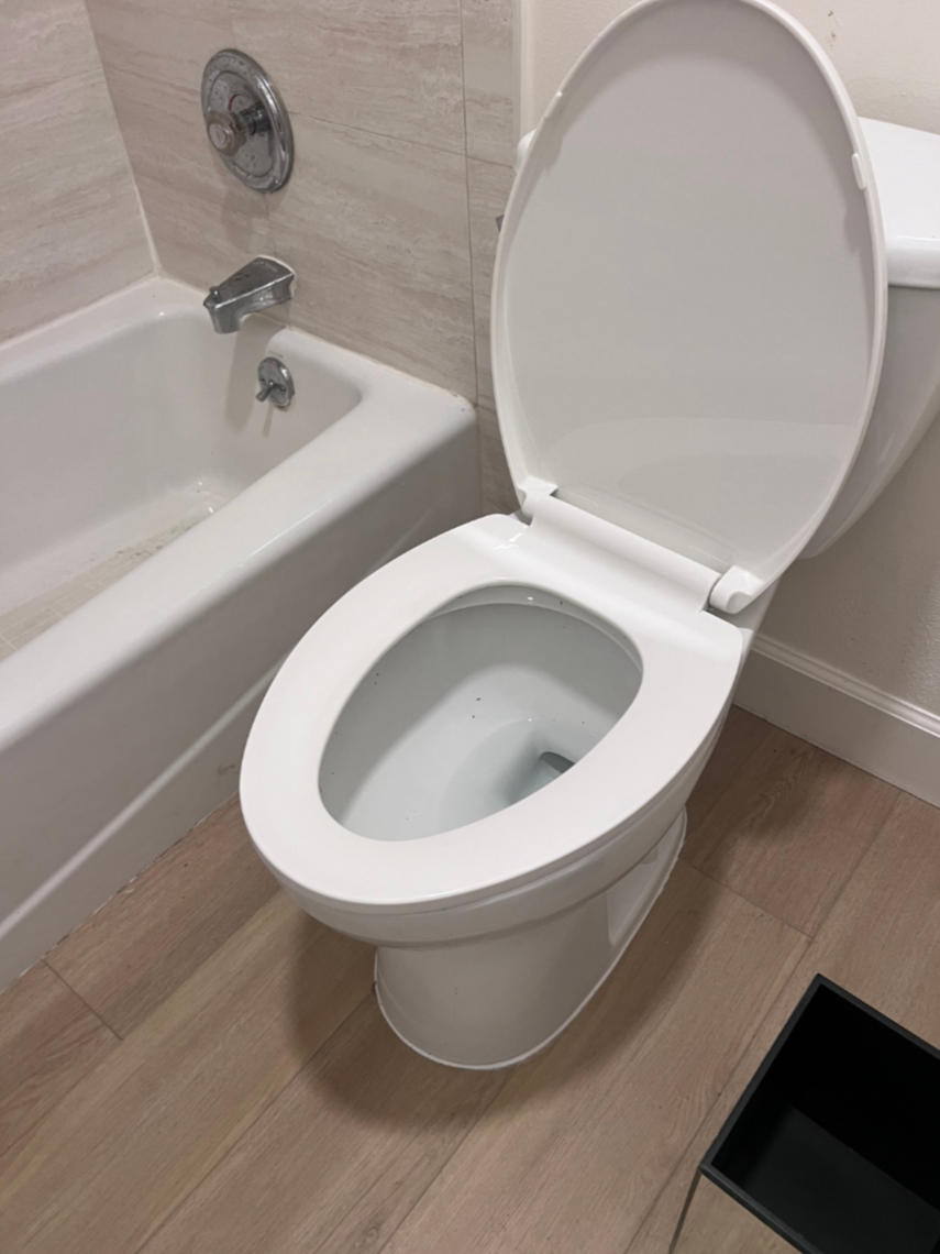Toilet with open lid next to a bathtub with a wall-mounted faucet and showerhead, in a bathroom with beige tile walls and wooden floor.