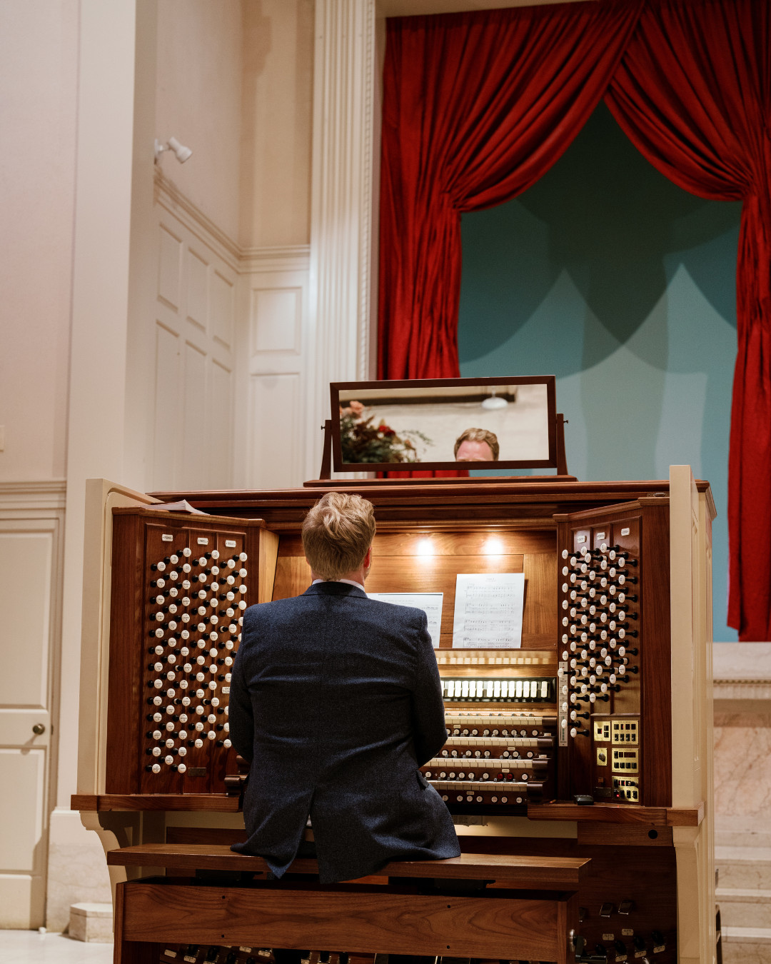 Wesley Hall, host of All the Stops podcast, sitting at an organ console in a church. His back is visible, along with the organ stops and manuals. There is a mirror on top of the console to view the audience.