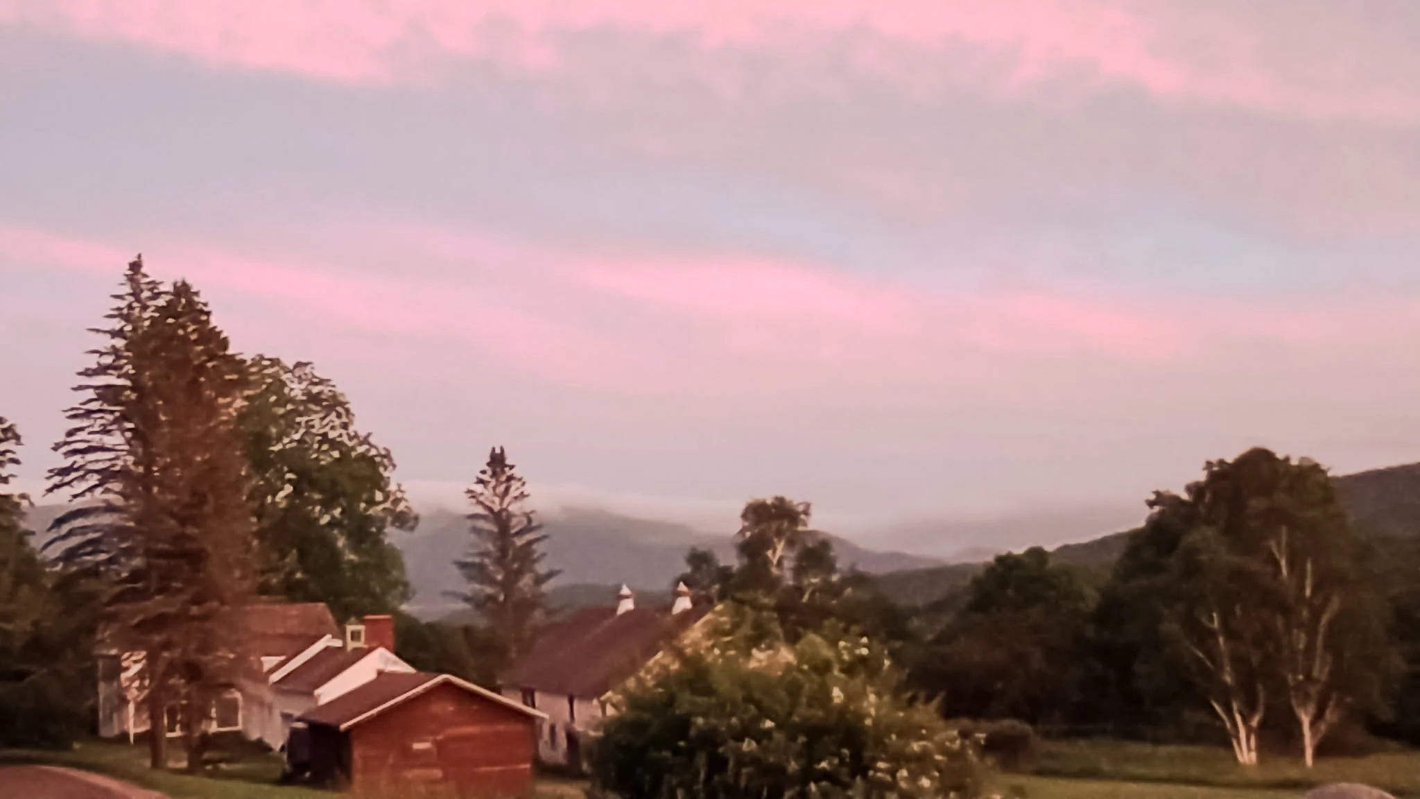 A peaceful landscape at sunset with houses, tall trees, rolling hills, and a purple-pink sky.