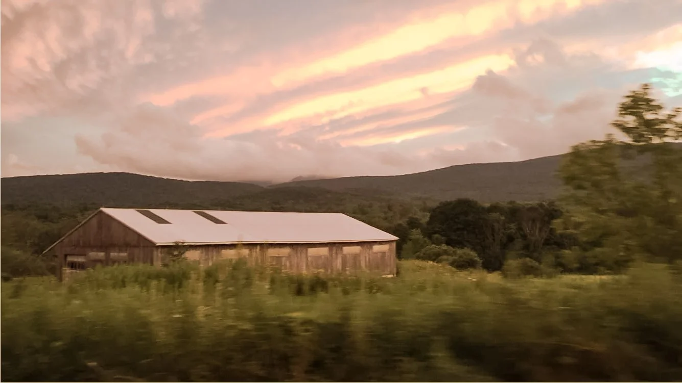 A rustic barn in a rural landscape during sunset, surrounded by green hills and trees.