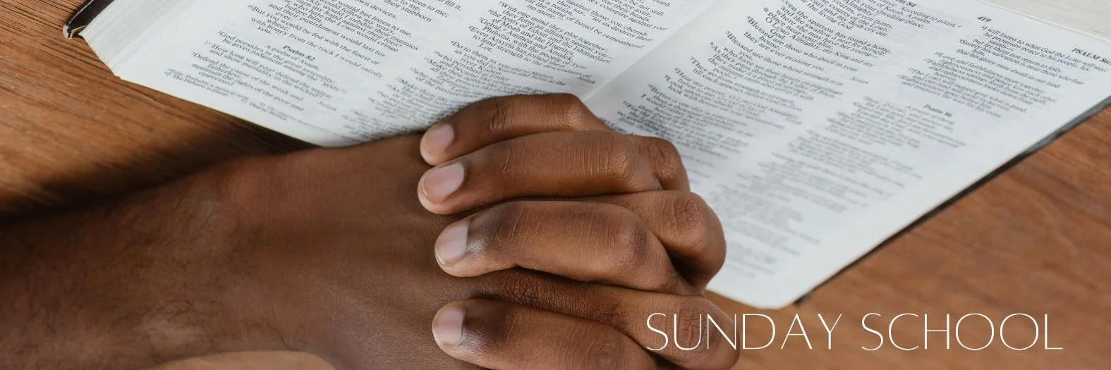 A person with dark skin praying with their hands clasped together on a wooden table, in front of an open Bible. The caption reads 'Sunday School'.