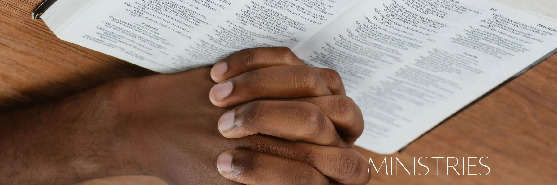 Two hands clasped together resting on an open Bible on a wooden surface, with the word 'MINISTRIES' in the bottom right corner.
