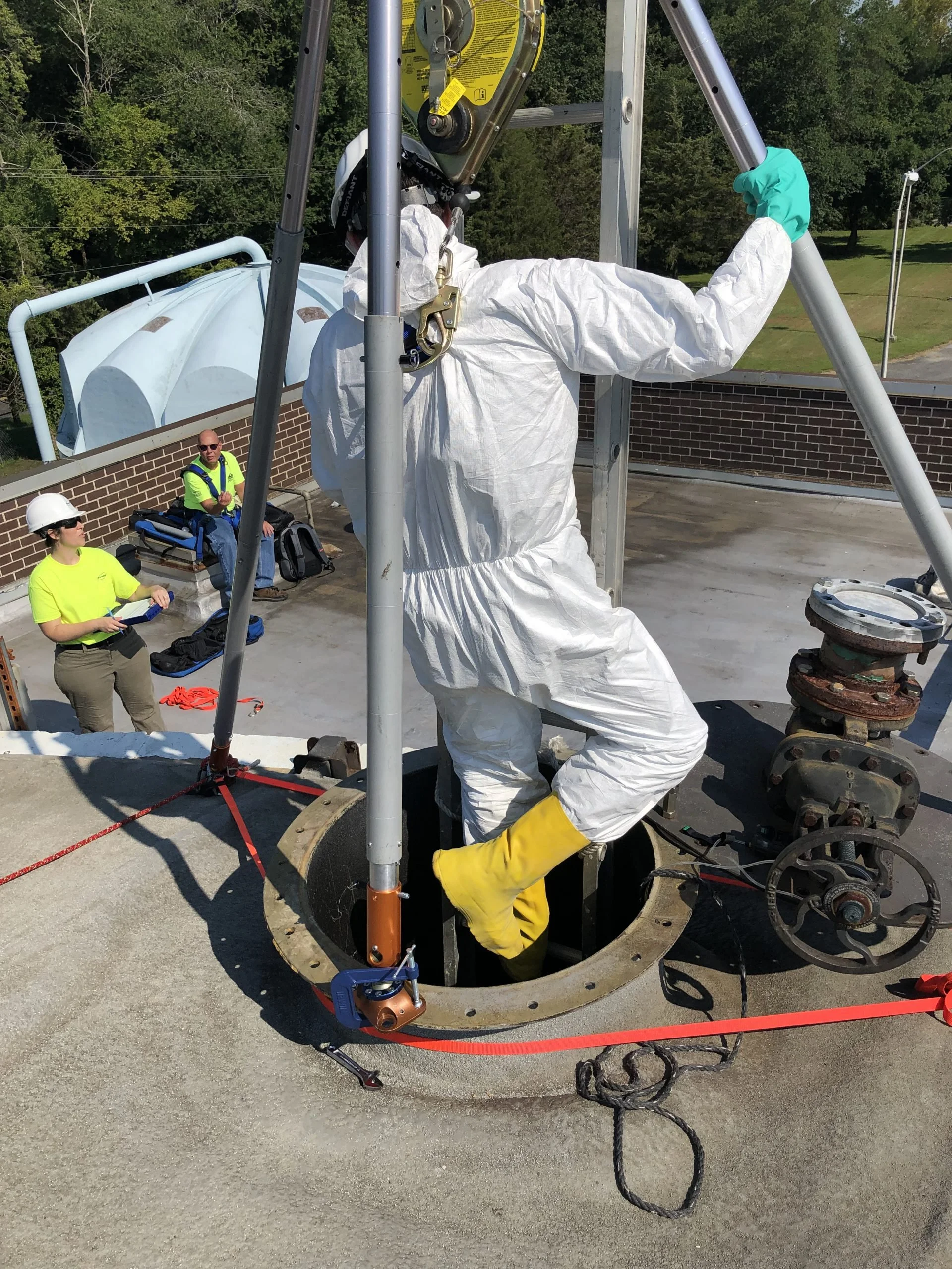 A worker in white protective gear and yellow boots is inside a manhole on a rooftop, working with tools surrounded by safety straps. Two other workers in yellow shirts and white hard hats are on the rooftop, observing and taking notes, with safety equipment nearby.
