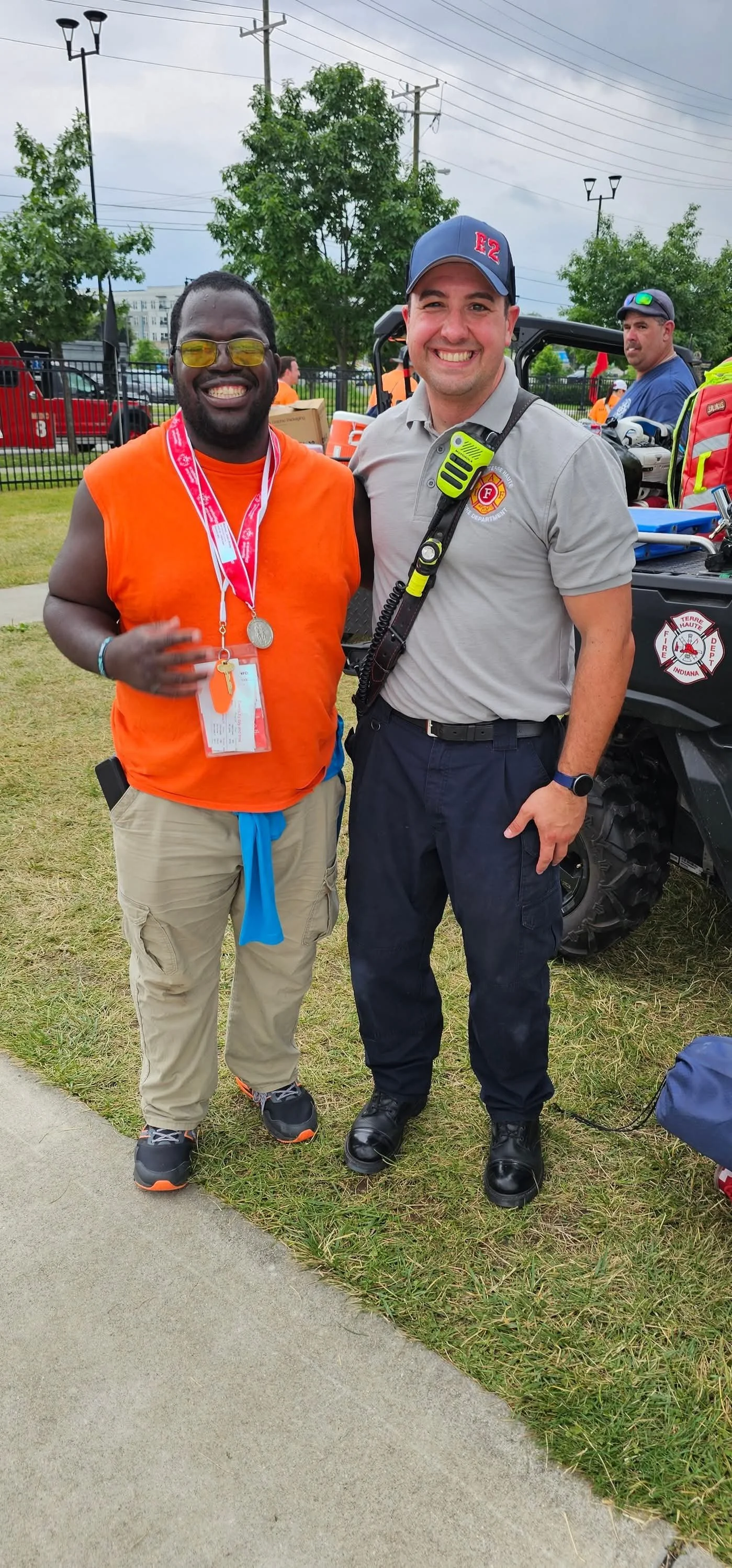 Two men standing outdoors at a community event, one wearing a bright orange sleeveless shirt with a medal and ID badge, the other in a gray uniform with a walkie-talkie, both smiling. A fire department vehicle and other people in orange are visible in the background.