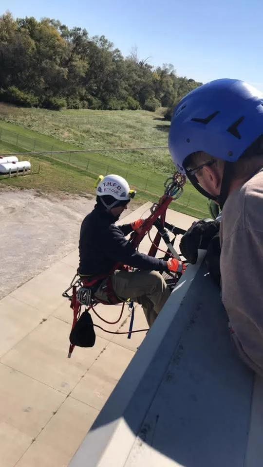 Two rescue workers in helmets and harnesses preparing on a rooftop for a rescue operation, with grassy fields and trees in the background.