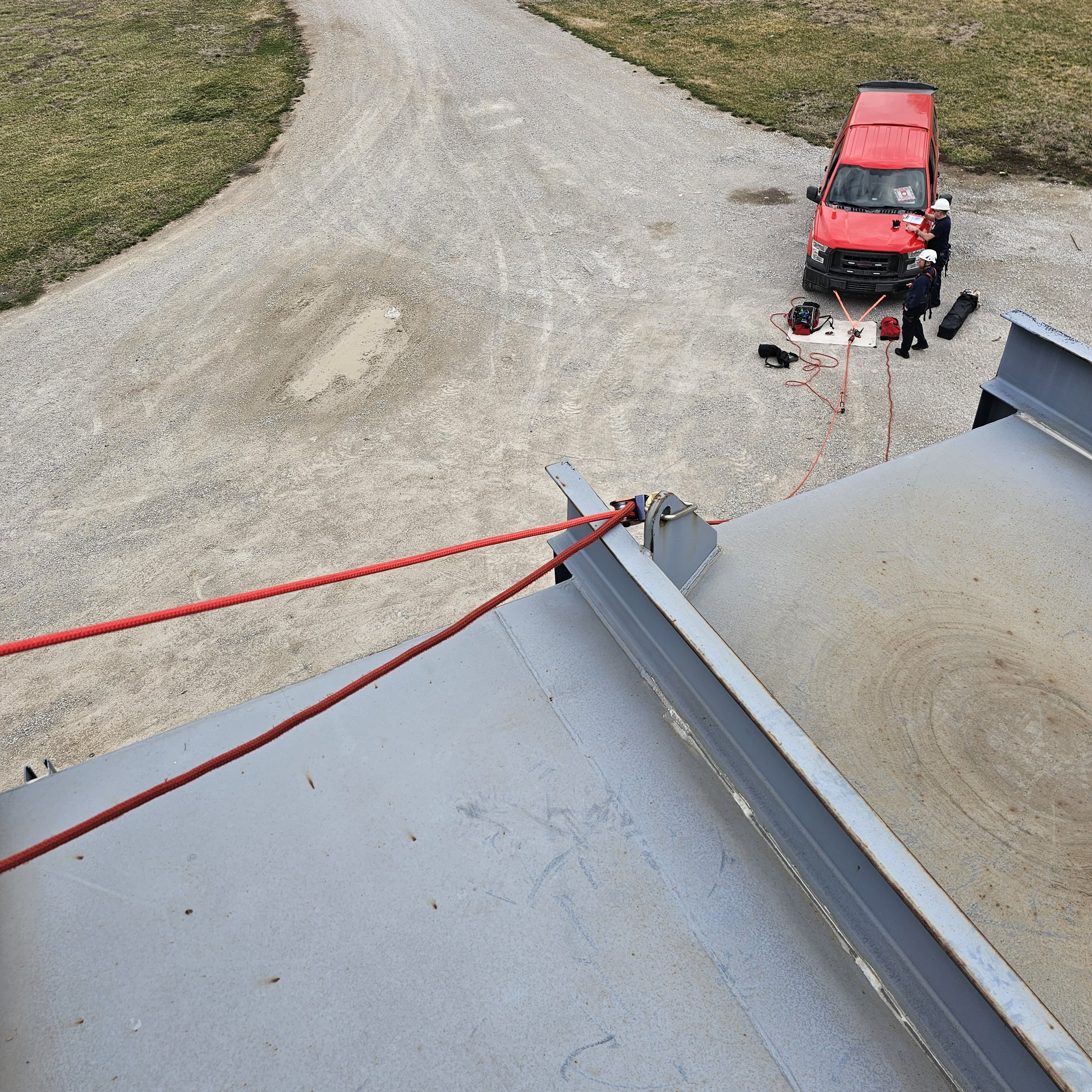 Aerial view of a gravel area with a red rescue vehicle and two emergency responders preparing equipment around it, with a large metal structure in the foreground.
