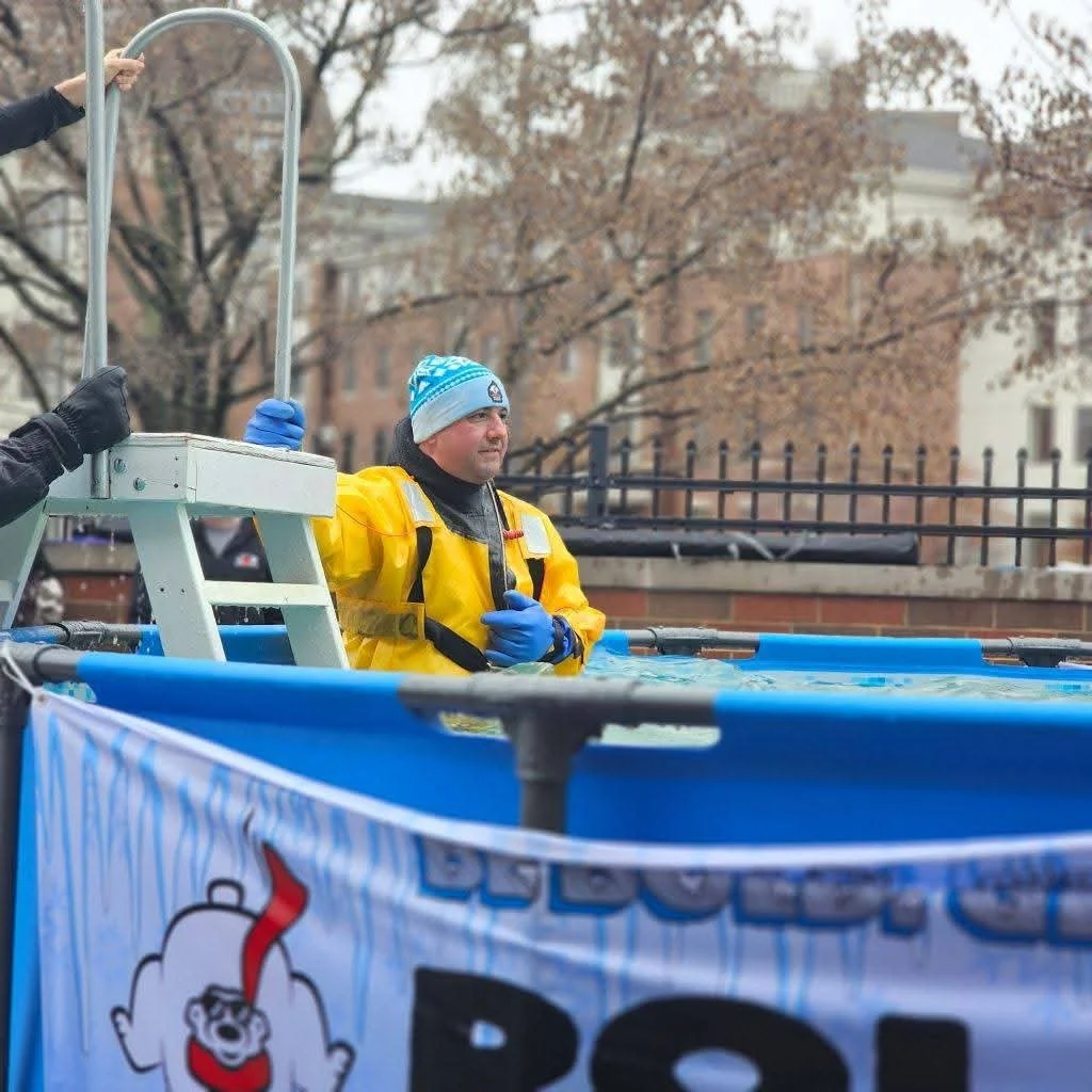 A man in a blue winter hat and yellow waterproof suit stands in a large blue tank, participating in a polar plunge event outdoors during fall or winter. There are leafless trees and a brick building in the background.