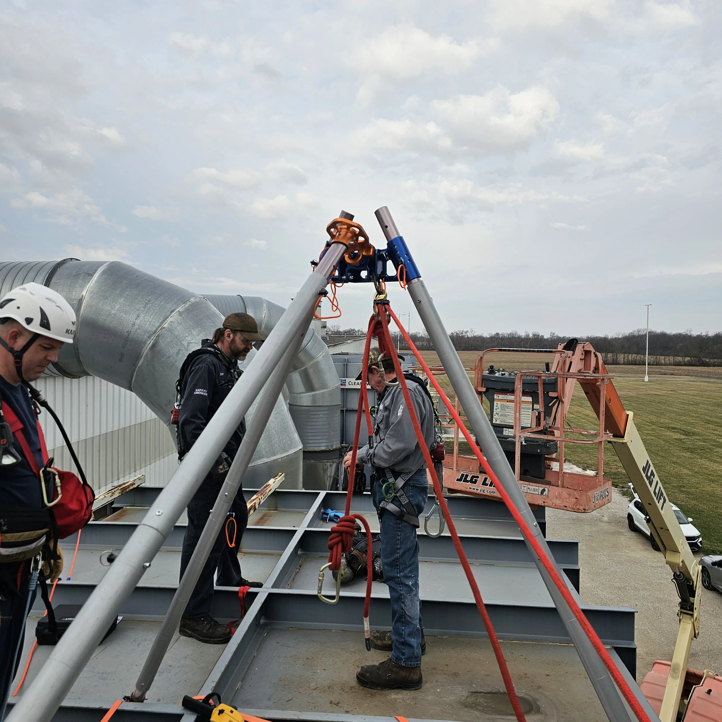 Workers installing or maintaining equipment on the roof of a building with safety harnesses, ropes, and tools, using a crane lift.