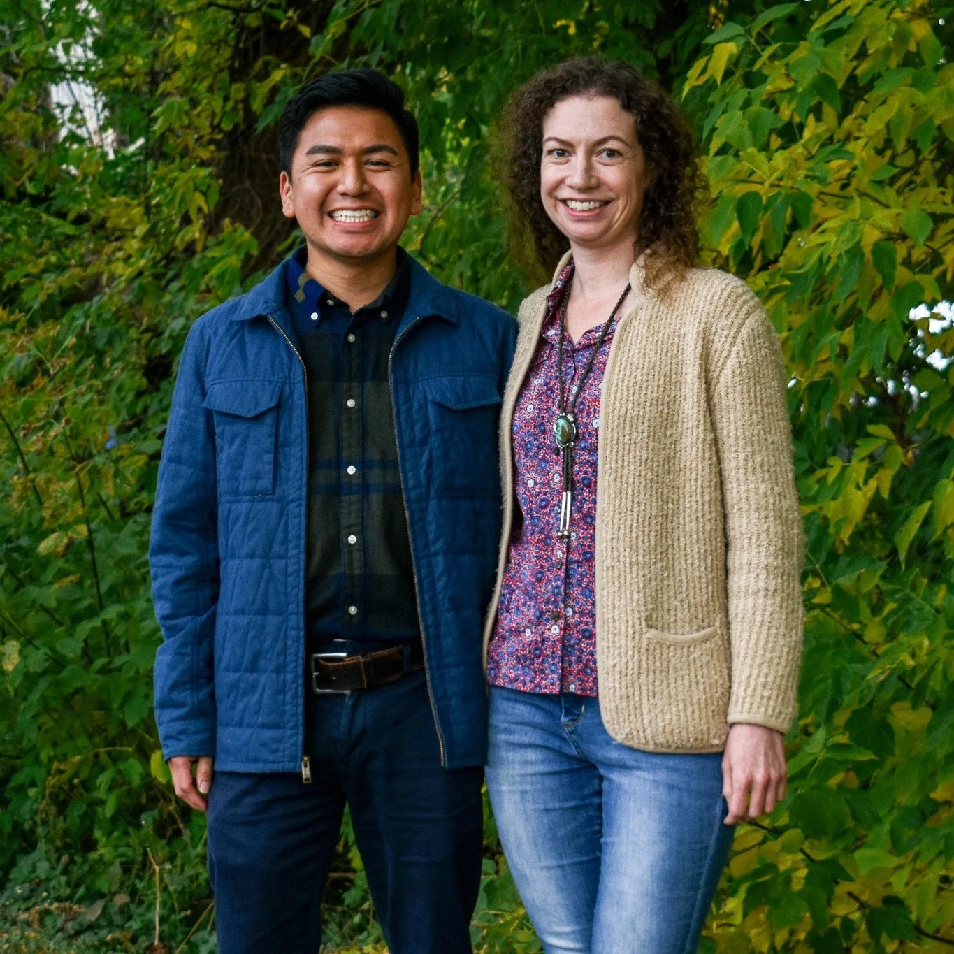 A young man and woman standing outdoors with green trees in the background, smiling at the camera.