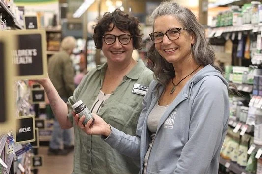 Two women smiling at a store aisle, holding small bottles of supplements or vitamins, with store shelves and other shoppers in the background.