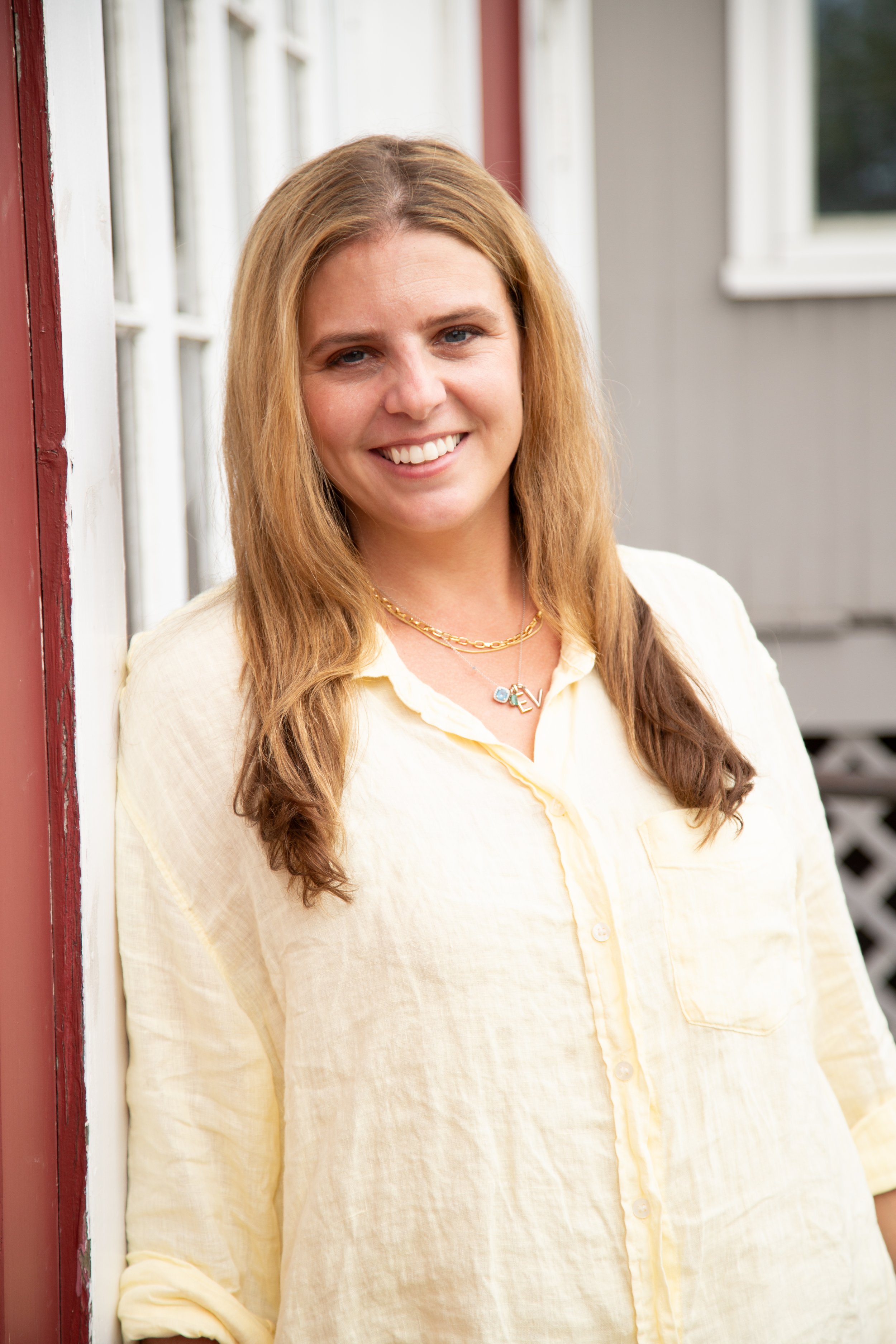 A headshot of Alexandra Nataloni, LMHC, therapist specializing in helping ambitious mothers with anxiety and burnout wearing a light yellow button-up shirt, standing outdoors next to a red and white building with windows.