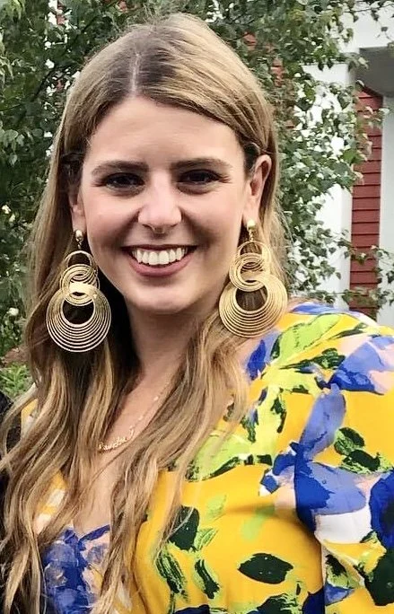 A young woman with long blonde hair wearing large gold hoop earrings and a colorful floral dress, smiling outdoors with trees and a house in the background.