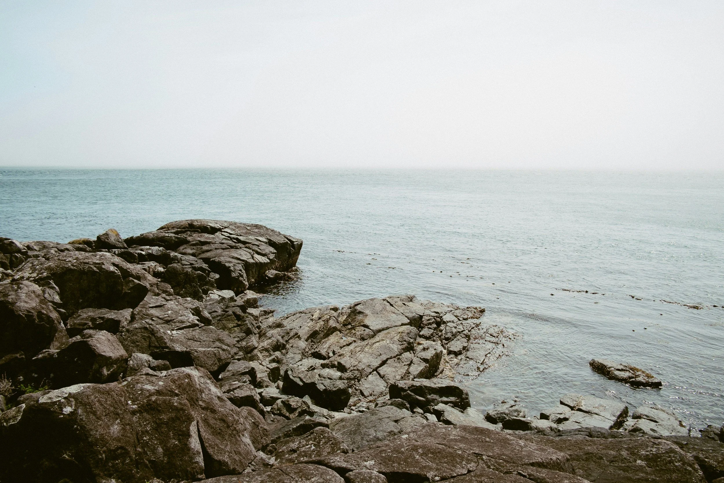 Rocky shoreline by the ocean with calm water and a hazy horizon in the distance.
