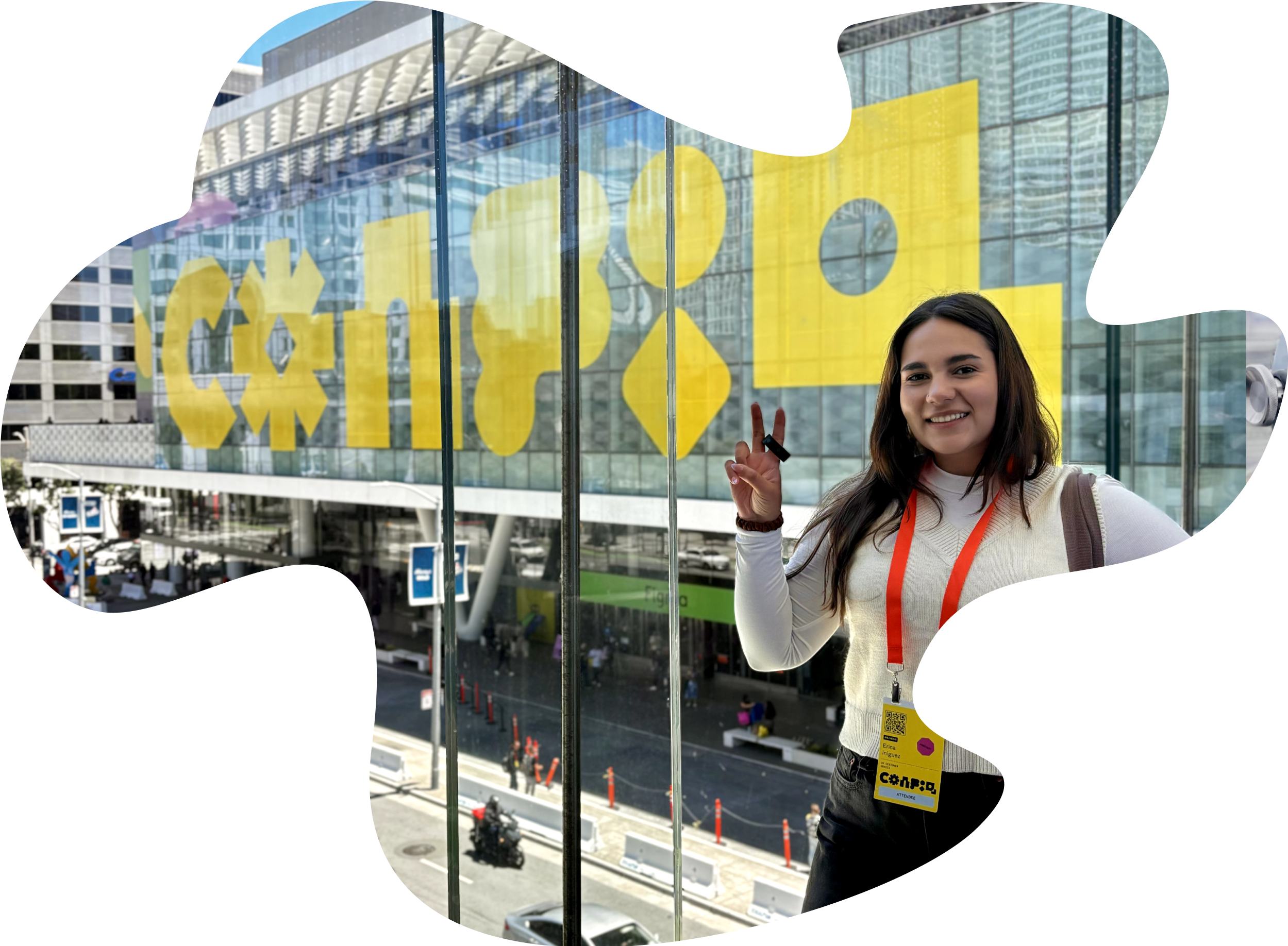 A young woman with dark brown hair smiling and making a peace sign with her right hand outside a modern glass building with yellow graphic and text on the window.