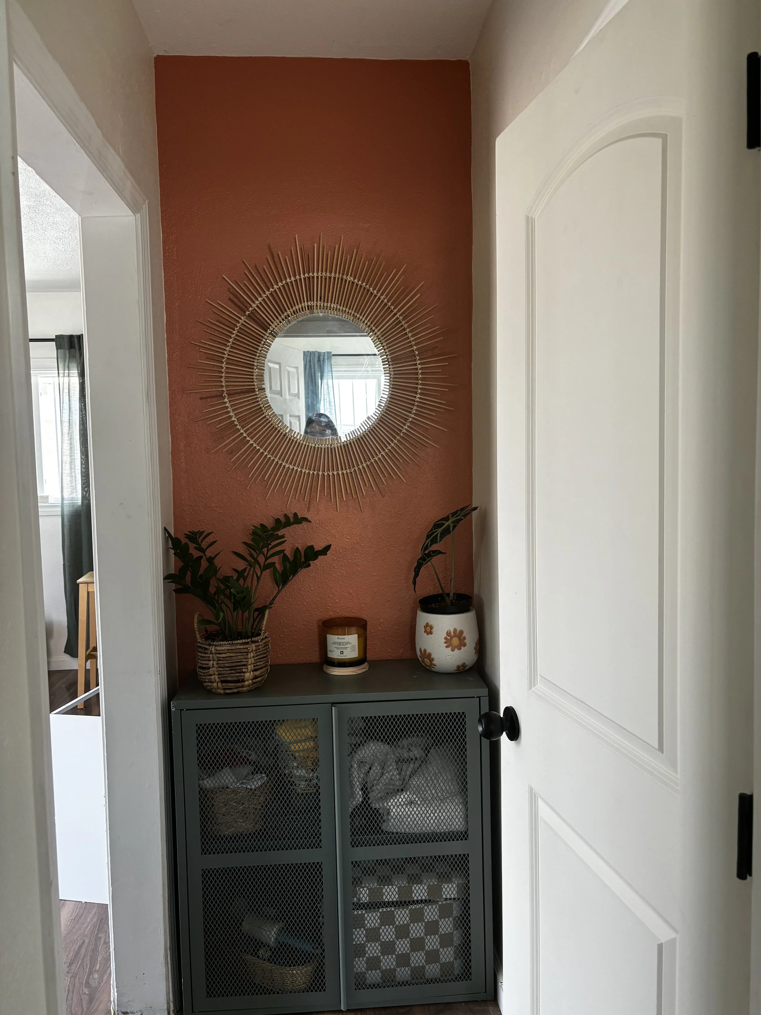 A hallway nook with a gold sunburst mirror on a terracotta wall, a gray cabinet with mesh doors and decorative baskets inside, and potted plants on top, including one with dark green leaves and another with large green leaves in a white flowered pot.